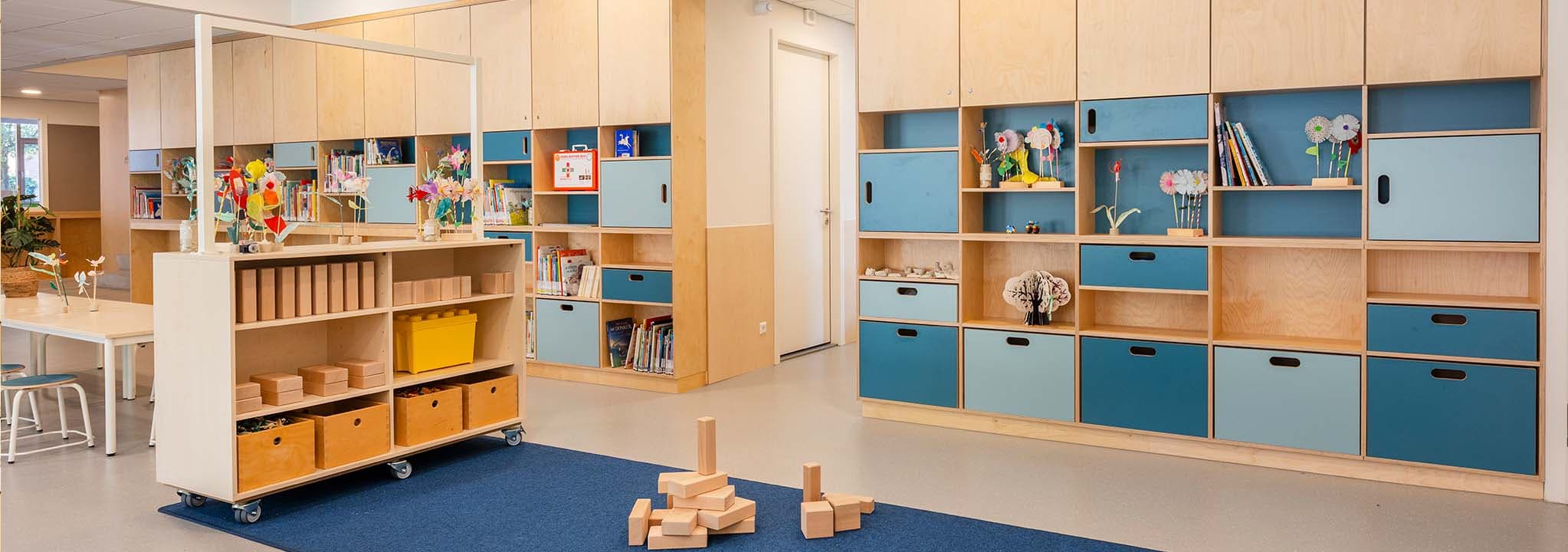 Kleurrijke groepsruimte op de opvang met houten opbergkasten tegen de wand met blauwe kastdeuren, en bouwblokken op een blauw vloerkleed.