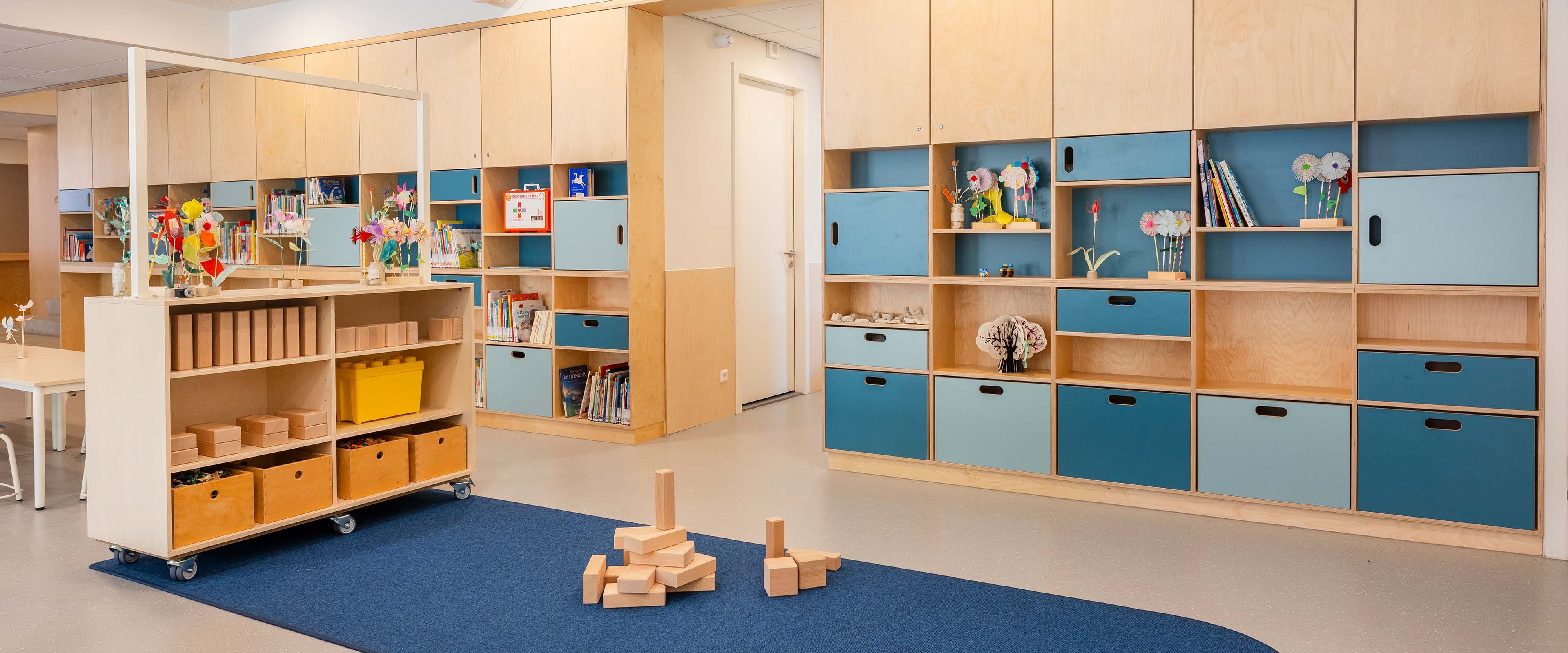 Kleurrijke groepsruimte op de opvang met houten opbergkasten tegen de wand met blauwe kastdeuren, en bouwblokken op een blauw vloerkleed.