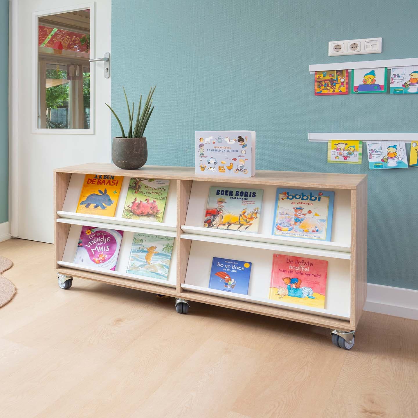 Houten boekenkast op wielen met kinderboeken, een plant en een puzzel in een lichte ruimte. De boeken zijn met de voorkant naar voren gepresenteerd.