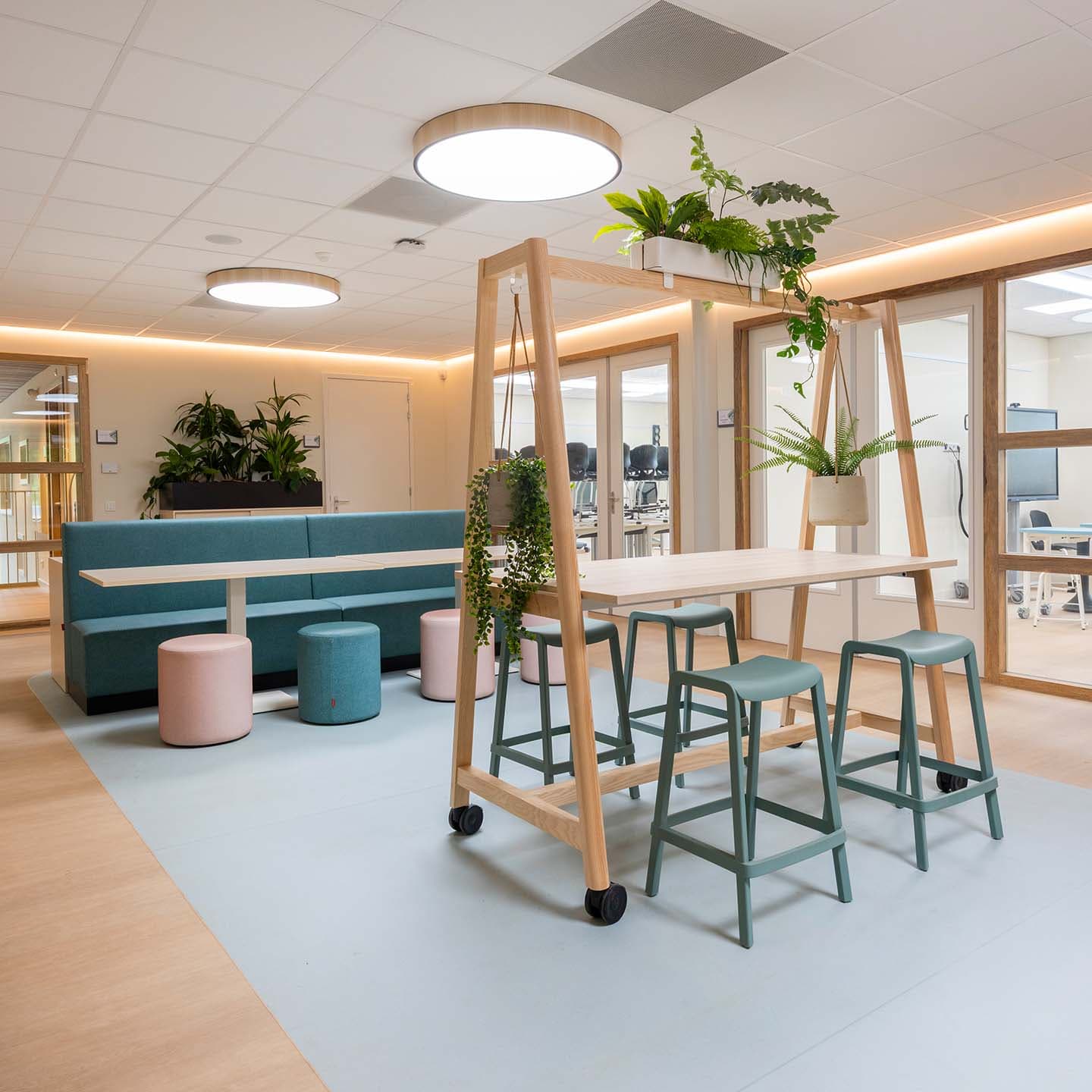 Open ruimte in de kinderopvang met een verrijdbare houten tafel, krukken, zitbanken en hangplanten in een lichte omgeving.