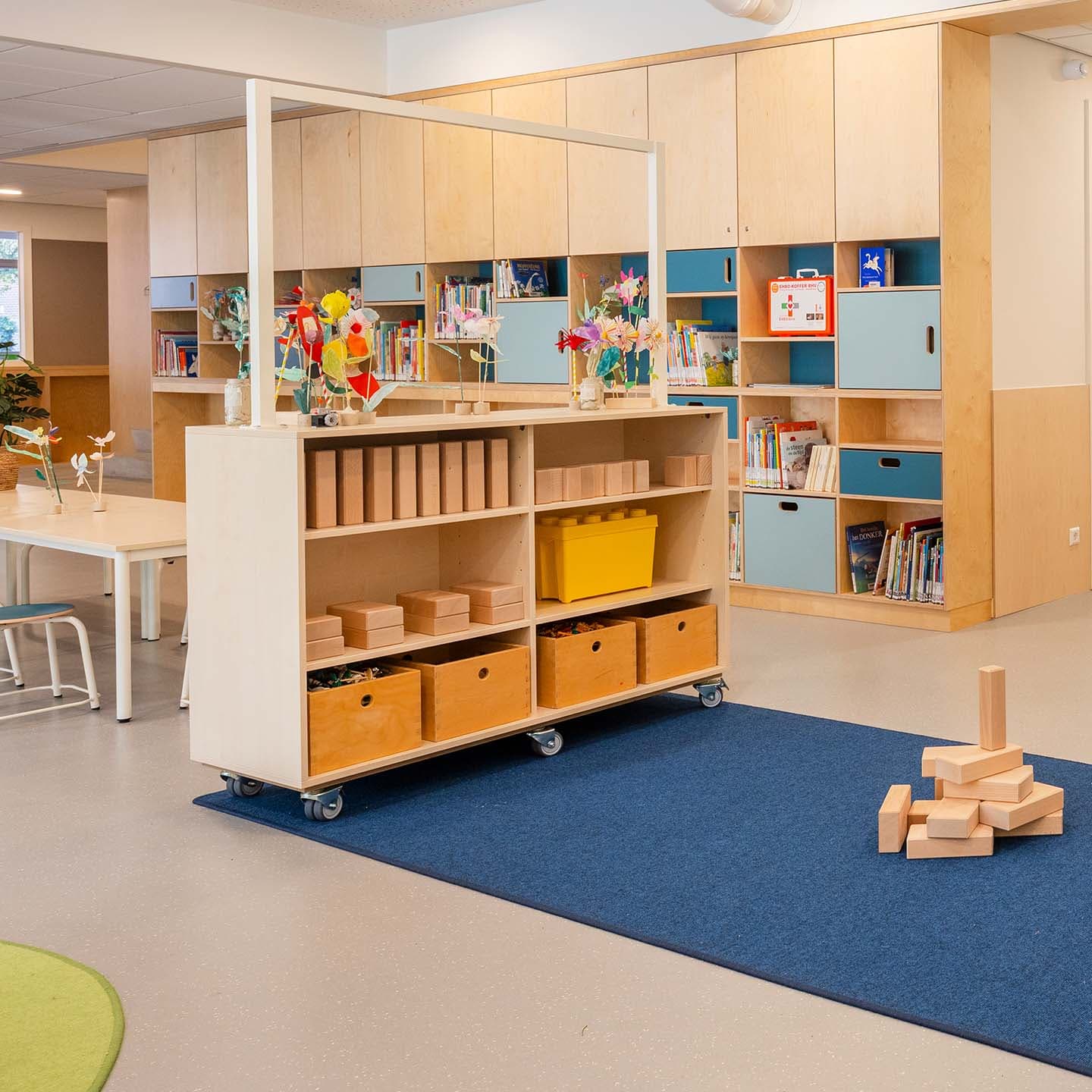 Een lichte groepsruimte in een kinderdagverblijf met kleurrijke planken, educatieve materialen, bloemen op een houten kar en houten blokken die zijn gestapeld op een blauw kleed.