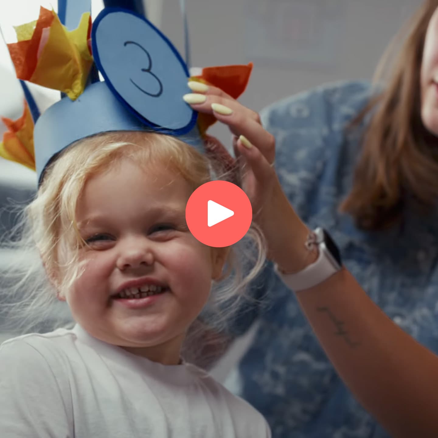 Child wearing a colorful paper crown with the number three, smiling happily as an adult adjusts it.