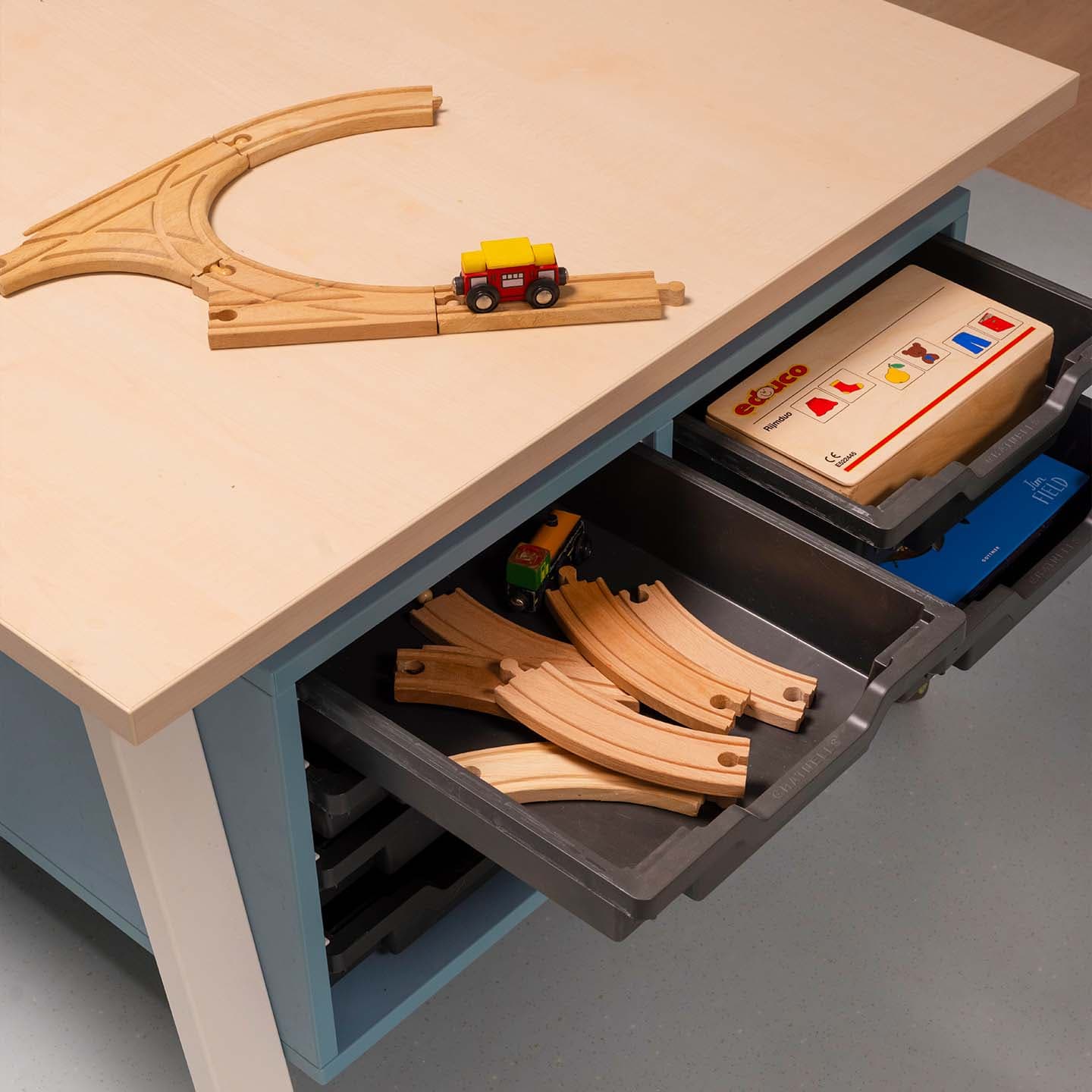 Houten treinrails en een speelgoedtrein op een opbergtafel met daaronder open lades gevuld met extra houten treinrails.