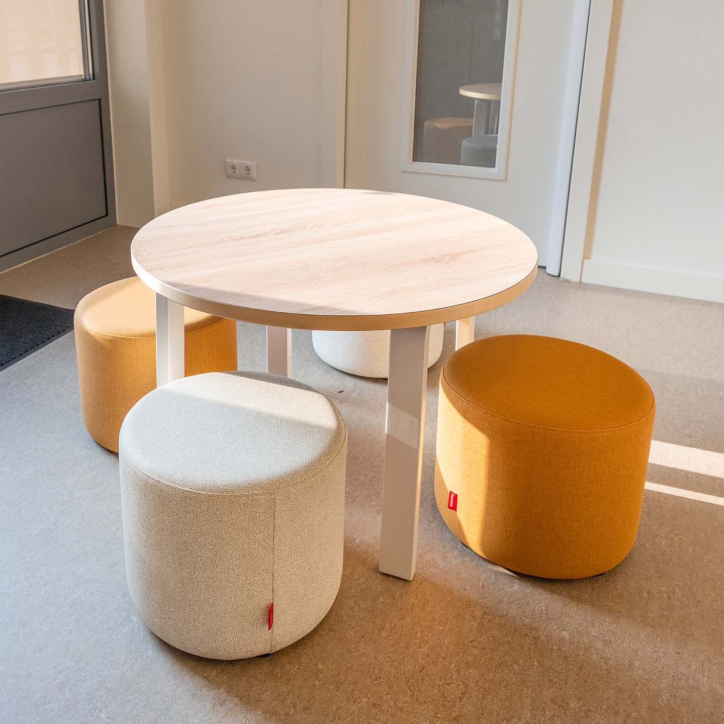 Ronde houten kindertafel met vier gestoffeerde poefjes in crèmekleur en okergeel in een lichte ruimte.
