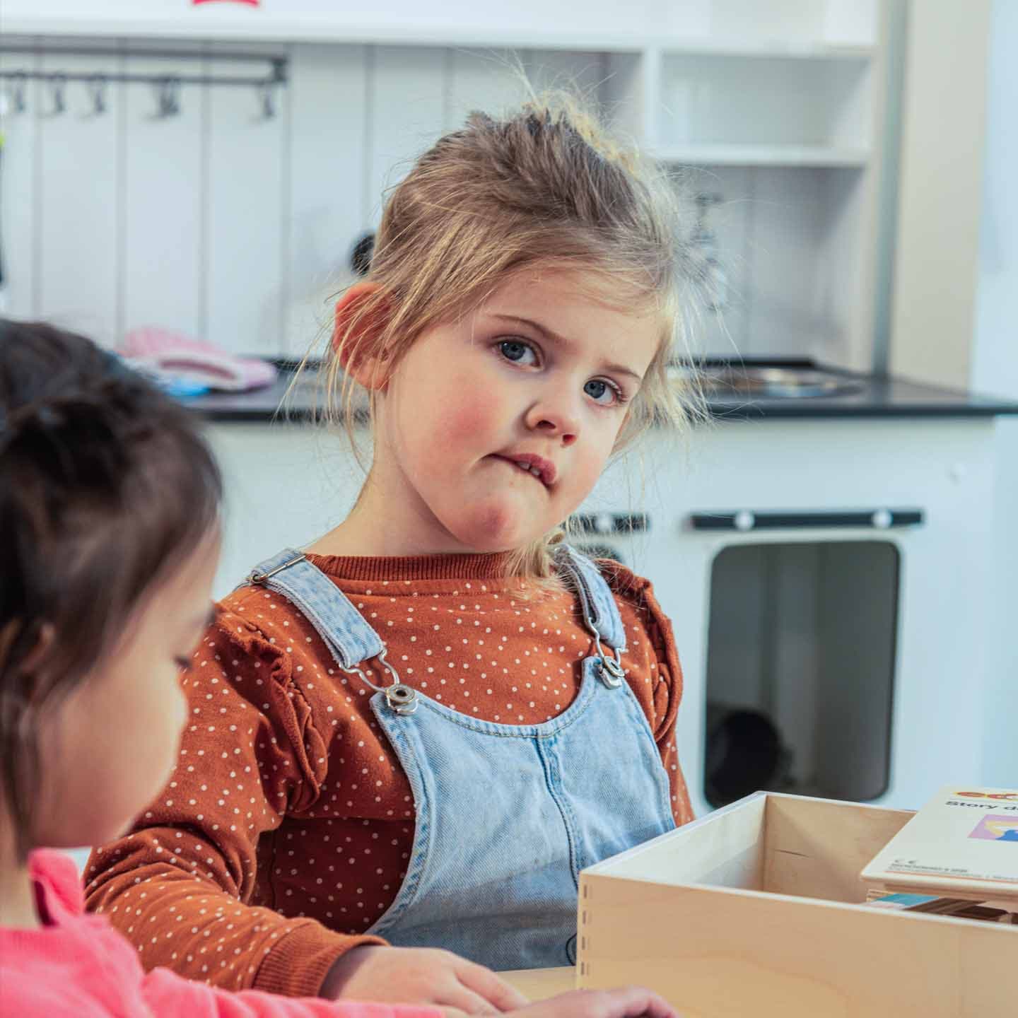Twee jonge meisjes spelen in een keukentje. Een van de meisjes, in een shirt met stippen en een spijkeroverall, kijkt nieuwsgierig in een houten doos.