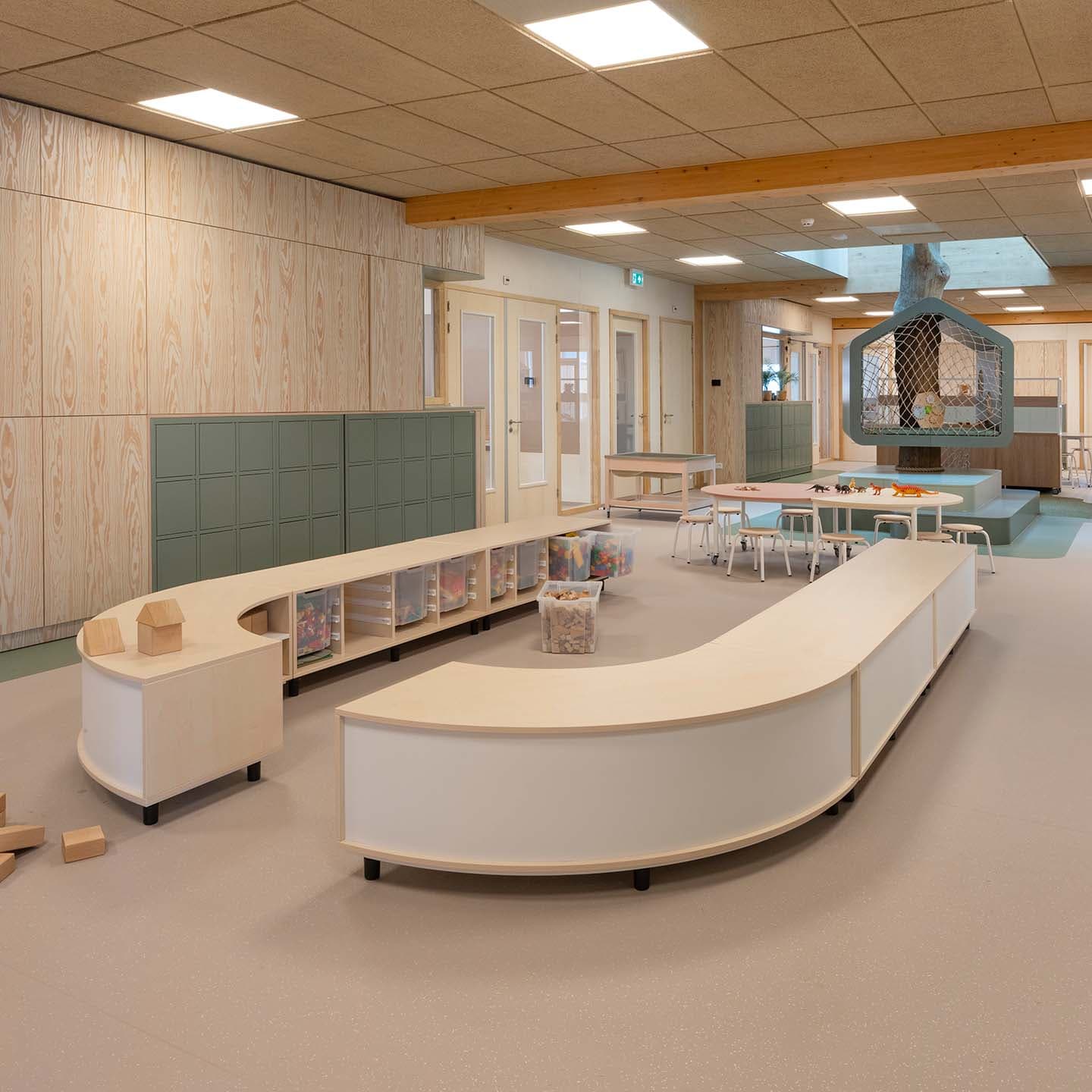Speelruimte in de kinderopvang met gebogen zitbanken, opbergvakken en houten bouwblokken. Natuurlijke materialen en rustige kleuren zorgen voor een warme uitstraling.