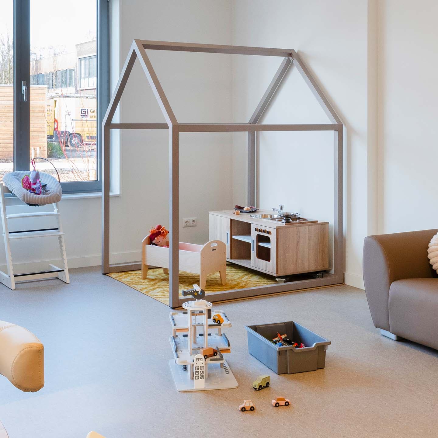 Speelruimte in de kinderopvang met een speelhuis van een stalen frame met een kinderkeuken, poppenbed en spelmateriaal op de vloer.