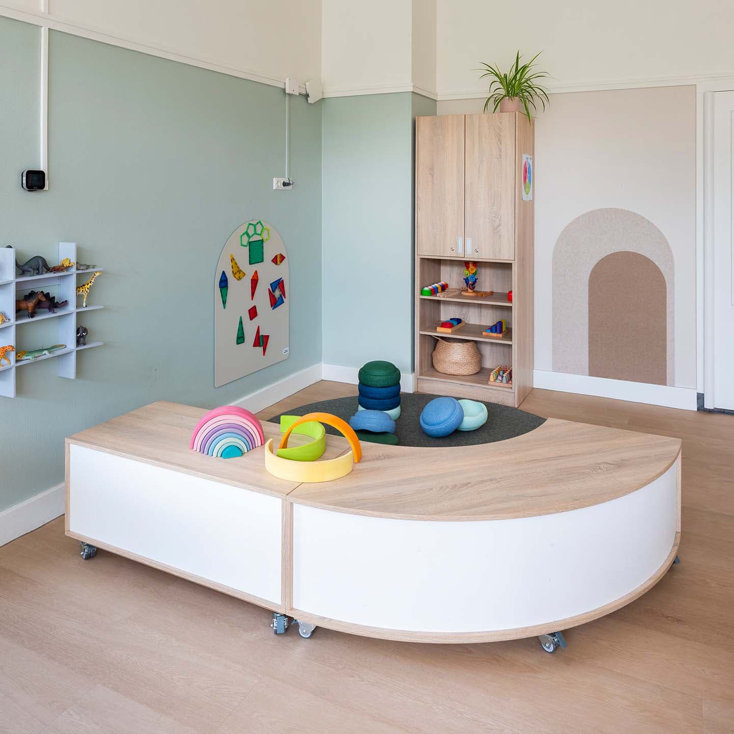 Speelruimte op de kinderopvang met lichtgroene wanden, houten ruimteverdelers, houten opbergkasten en een vloerkleed. Op al het meubilair ligt gekleurd open-ended speelgoed.