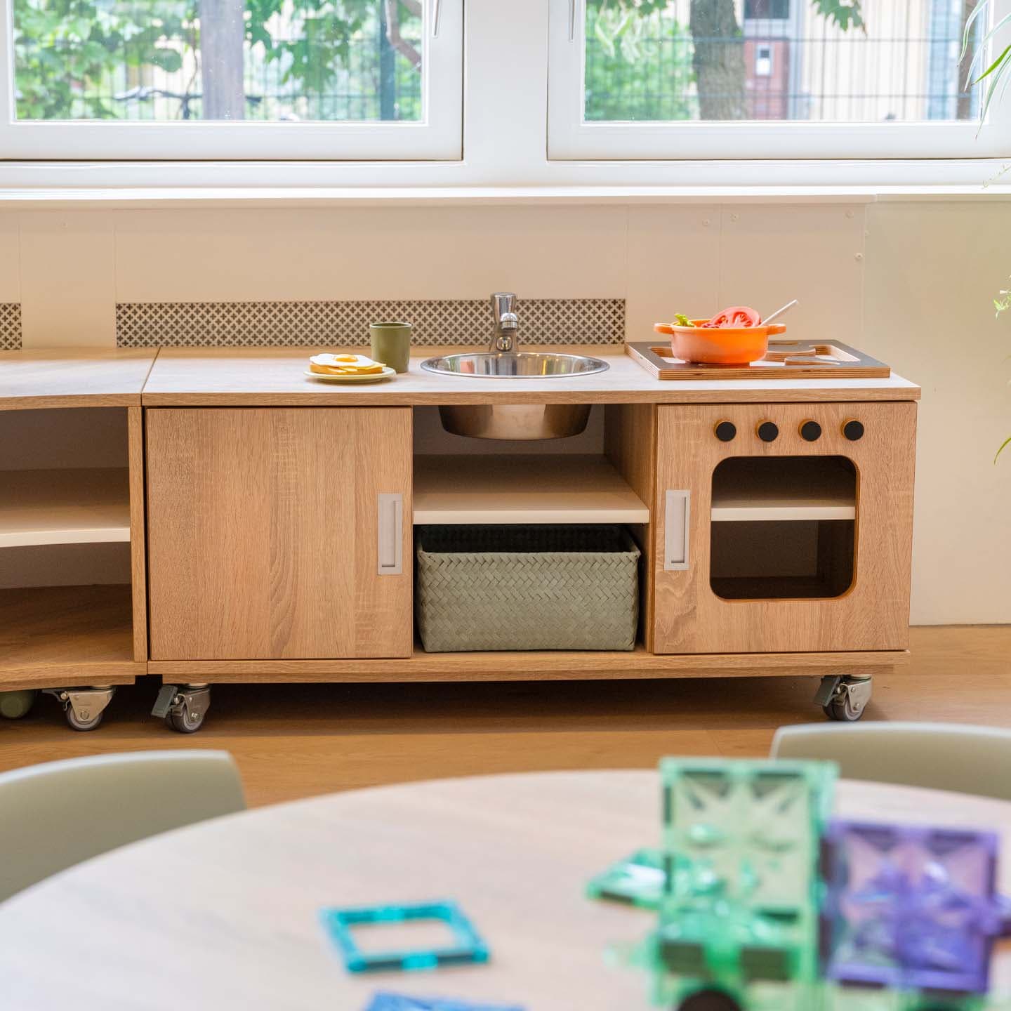 Kinderkeuken met houten kastjes, een spoelbak en een speelfornuis, met een ronde kindertafel en kleurrijke bouwblokken op de voorgrond.