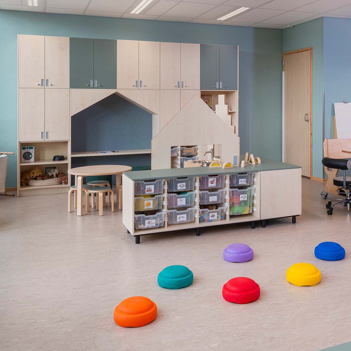 Een kleurrijke en overzichtelijke groepsruimte in een kinderdagverblijf met opbergbakken, ronde krukjes, een kleine tafel en een speelhoek in de vorm van een huis.