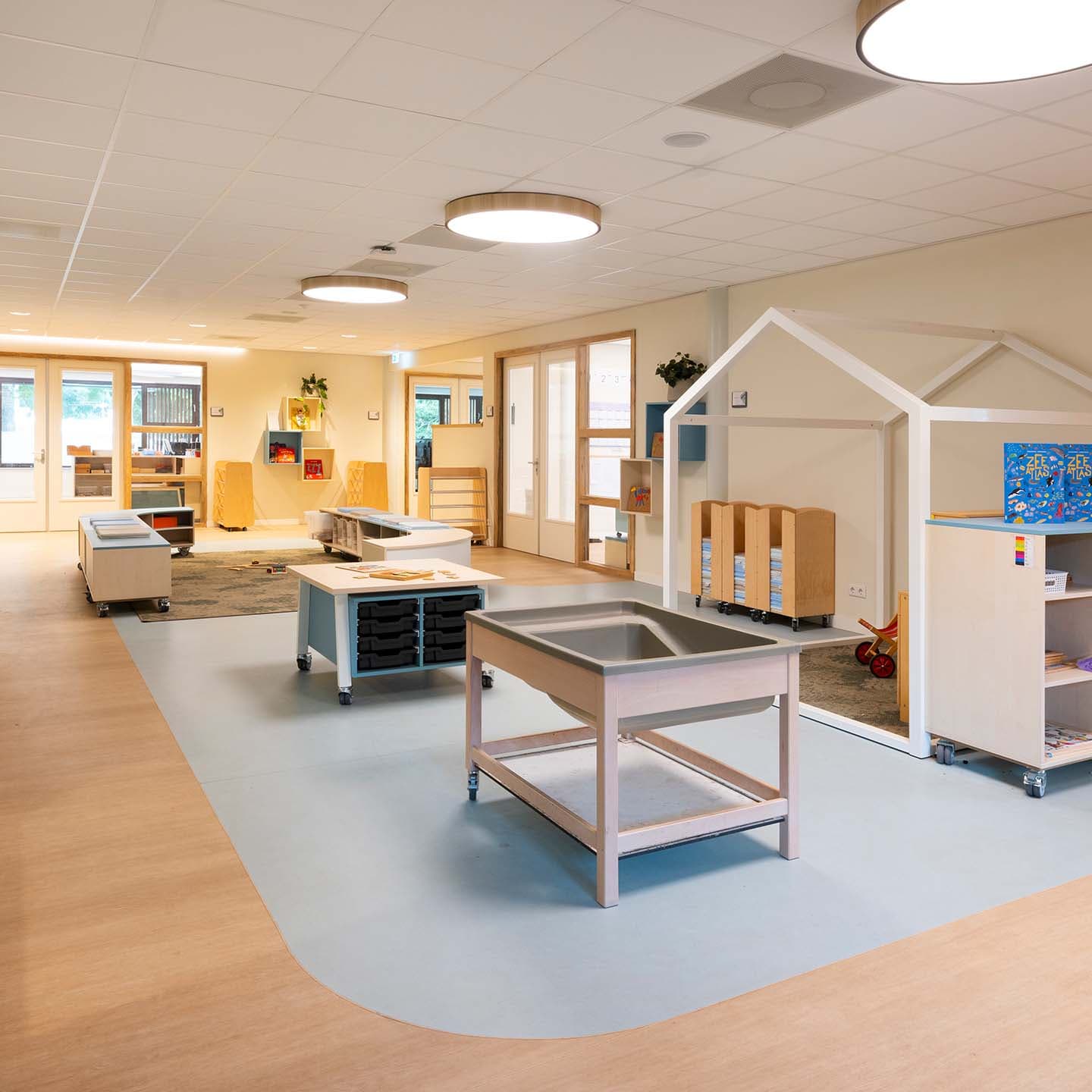 Ruime en open groepsruimte in de kinderopvang met verschillende speelplekken, een speelhuisje, opbergruimte op kinderhoogte en verrijdbare meubels voor een flexibele inrichting.