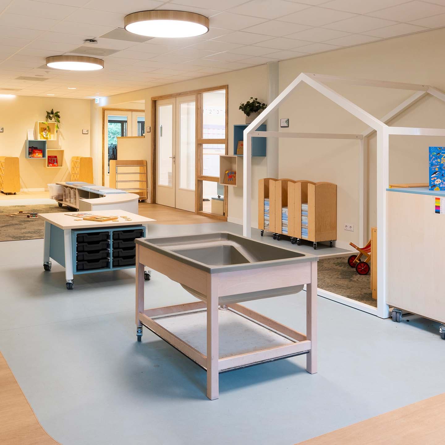 Een lichte en ruimtelijke groepsruimte in een kinderdagverblijf met tafels, opbergmeubels, een speelhuisconstructie en kinderspeelgoed, wat zorgt voor een uitnodigende leeromgeving.