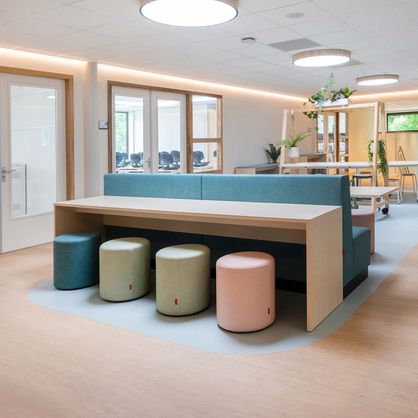 Centrale ruimte op de basisschool met een lange houten tafel, pastelkleurige poefjes die ook weer terugkomen in een bank.