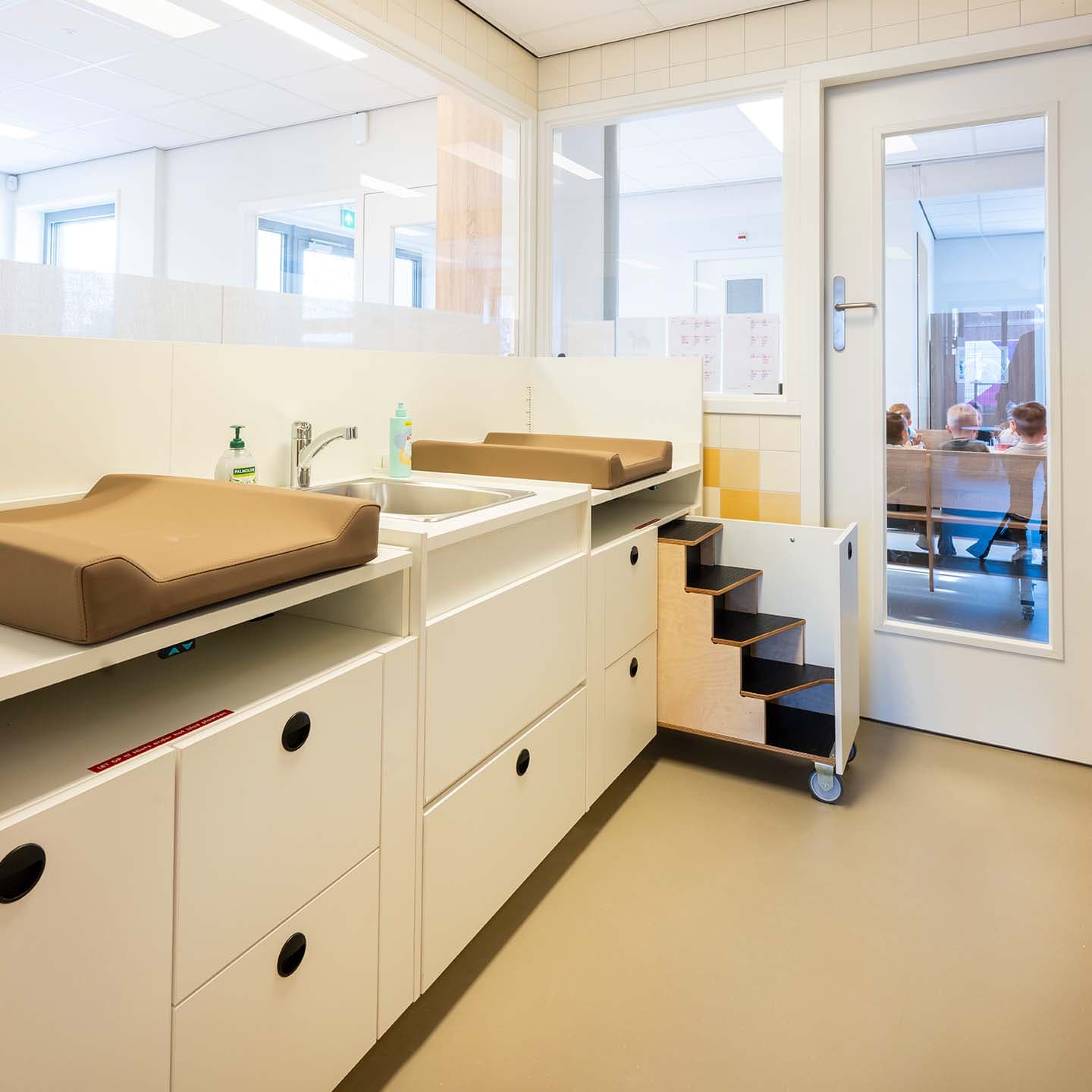 Verschoonruimte in de kinderopvang met twee verschoonplekken, een wastafel, opbergkasten en een deur naar de groepsruimte.
