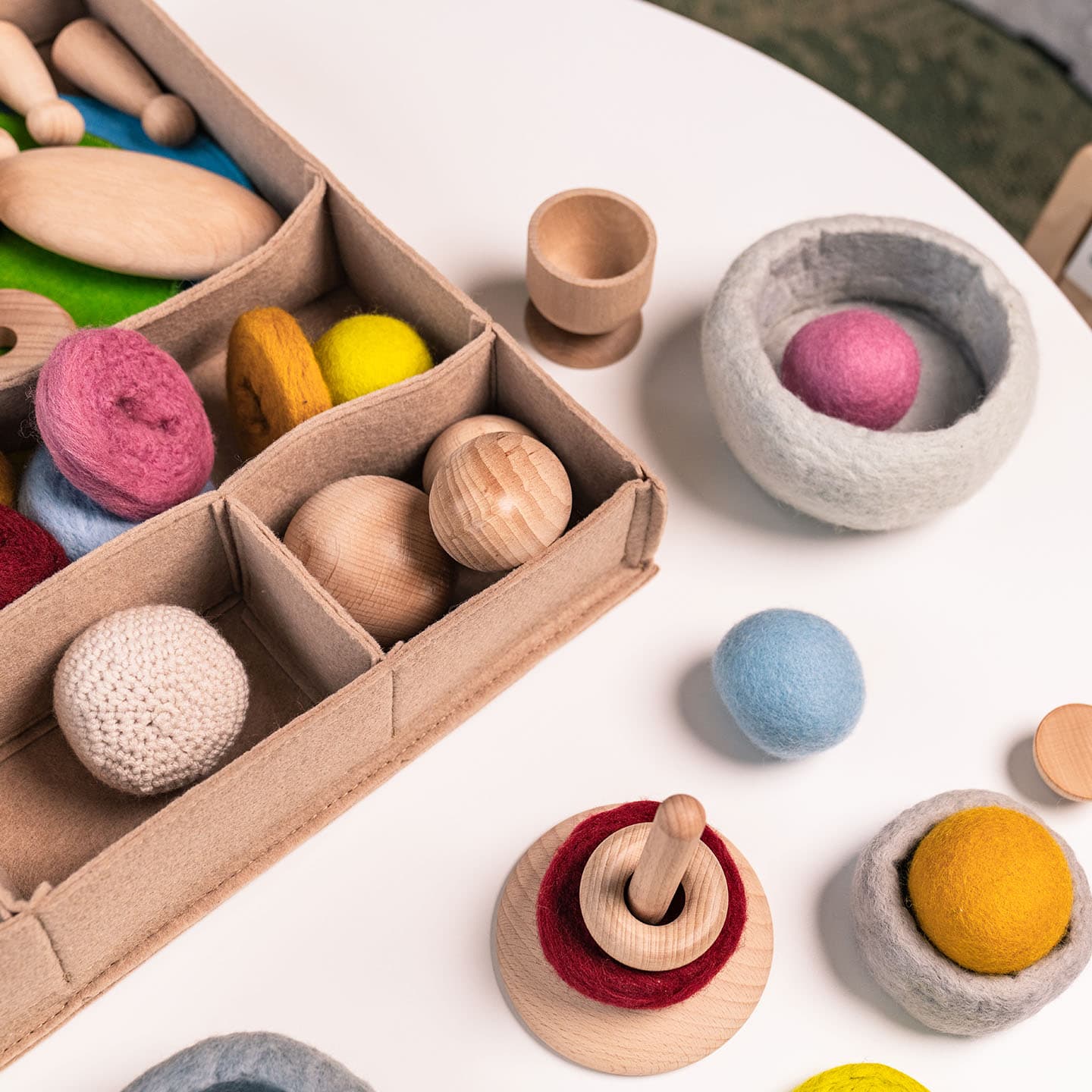 Houten en vilten speelgoed, waaronder ballen en stapelblokken, liggen uitgestald op een witte tafel met een doos met vakjes. Op de achtergrond zijn borden en bestek wazig te zien.