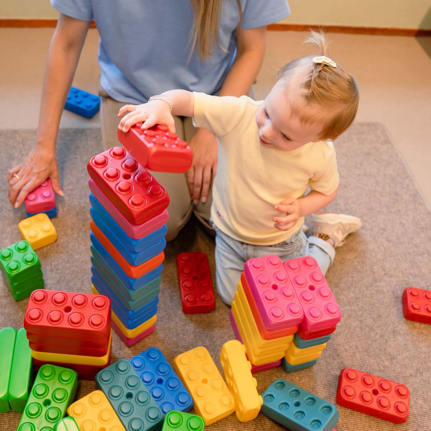 Een peuter speelt met kleurrijke bouwblokken op een vloer met tapijt, en stapelt ze op onder begeleiding van een pedagogisch professional die er vlakbij zit.