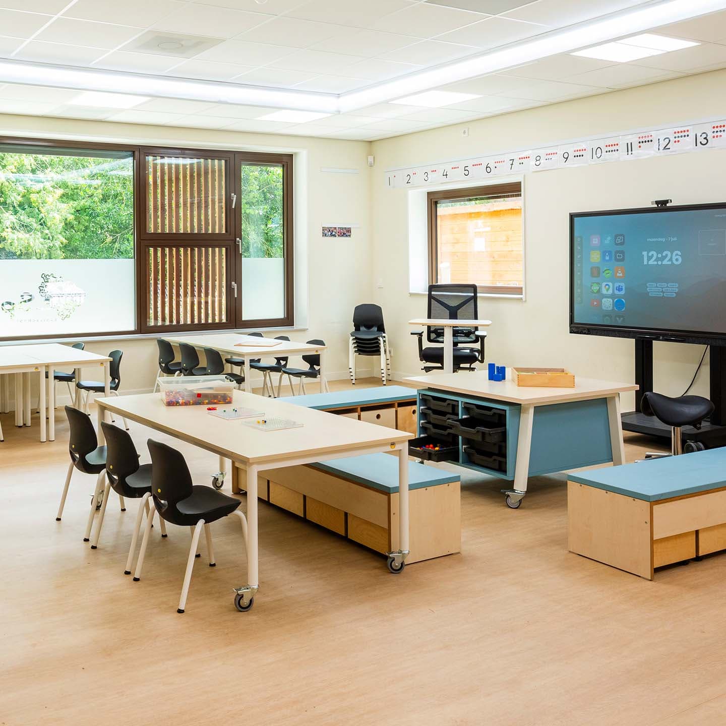 Modern klaslokaal met tafels, stoelen, een interactief scherm en grote ramen. Licht en overzichtelijk ingericht met educatieve materialen aan de wanden.