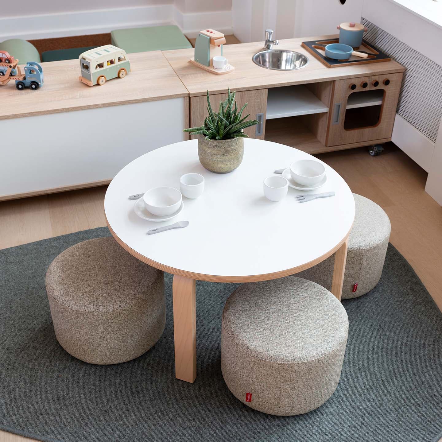 Ronde houten kindertafel met poefjes, een speeltheeservies en op de achtergrond een kinderkeuken met houten keukengerei.