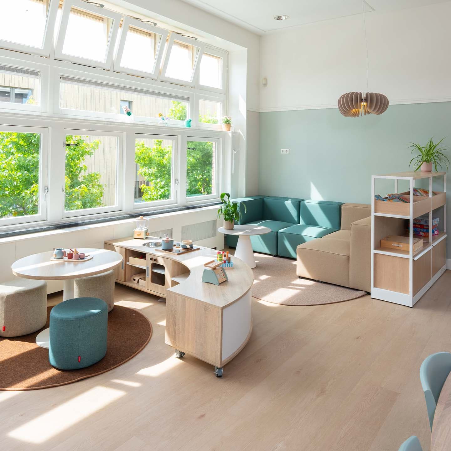 Een lichte kinderspeelkamer met ronde tafels, krukjes en een turquoise bank bij grote ramen. Op planken staan speelgoed en een plant siert de hoek.