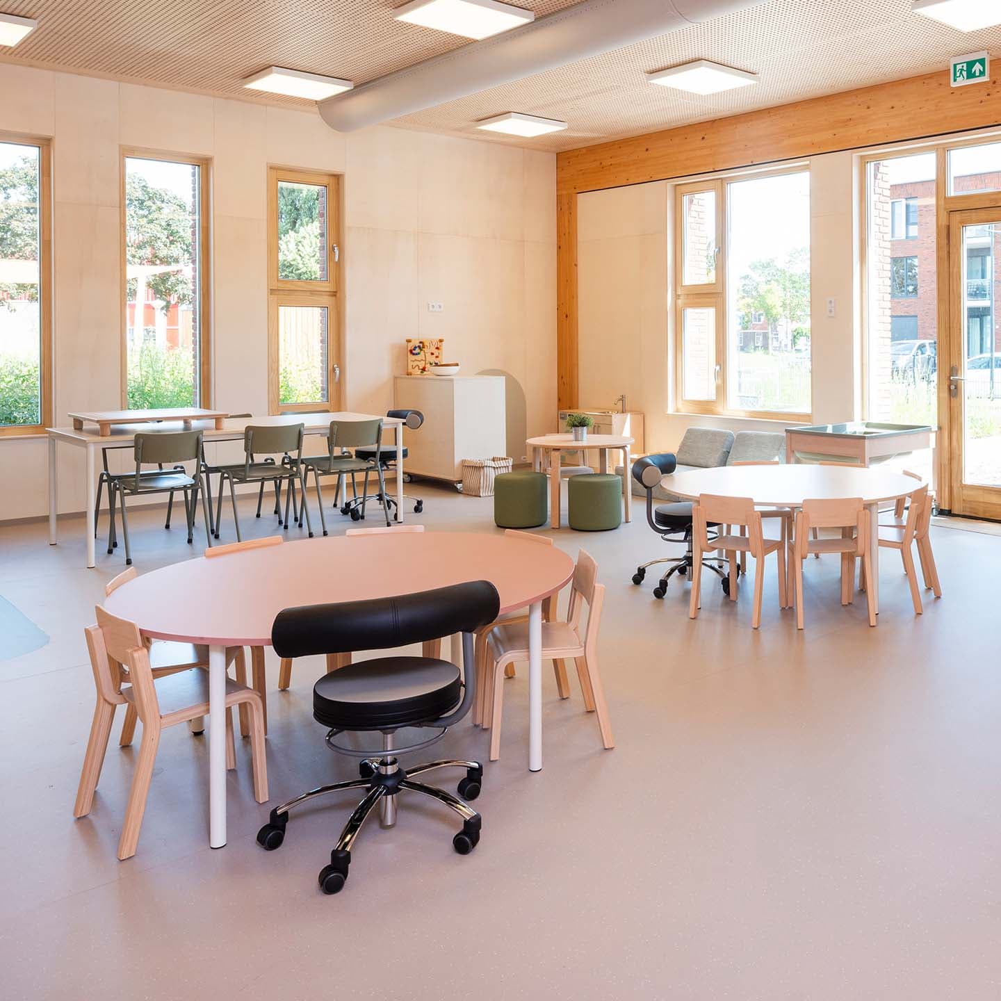 Een lichte, ruime groepsruimte in een kinderdagverblijf met ronde tafels, houten stoelen, een verrijdbare stoel en grote ramen die natuurlijk licht binnenlaten.