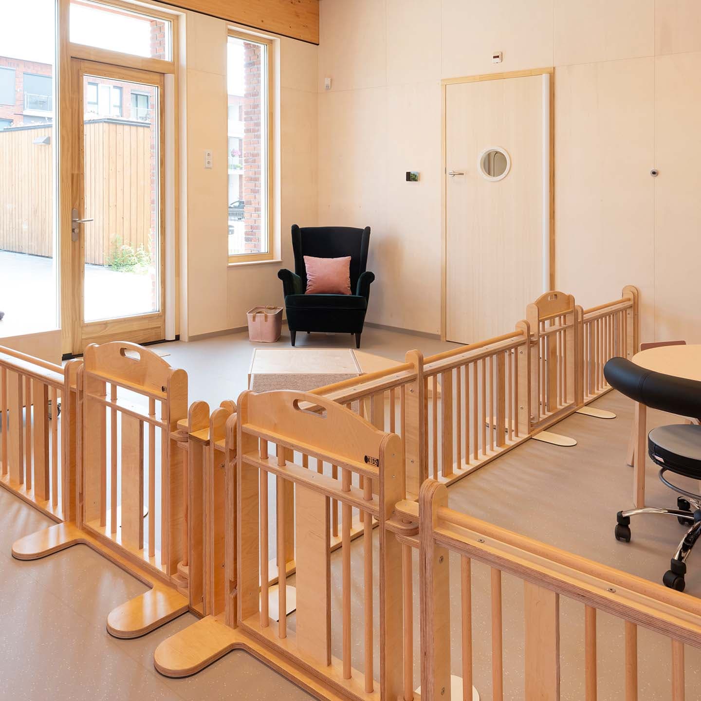 Lichte ruimte in de kinderopvang met houten grondbox, een ronde tafel, een fauteuil en grote ramen met veel daglicht.
