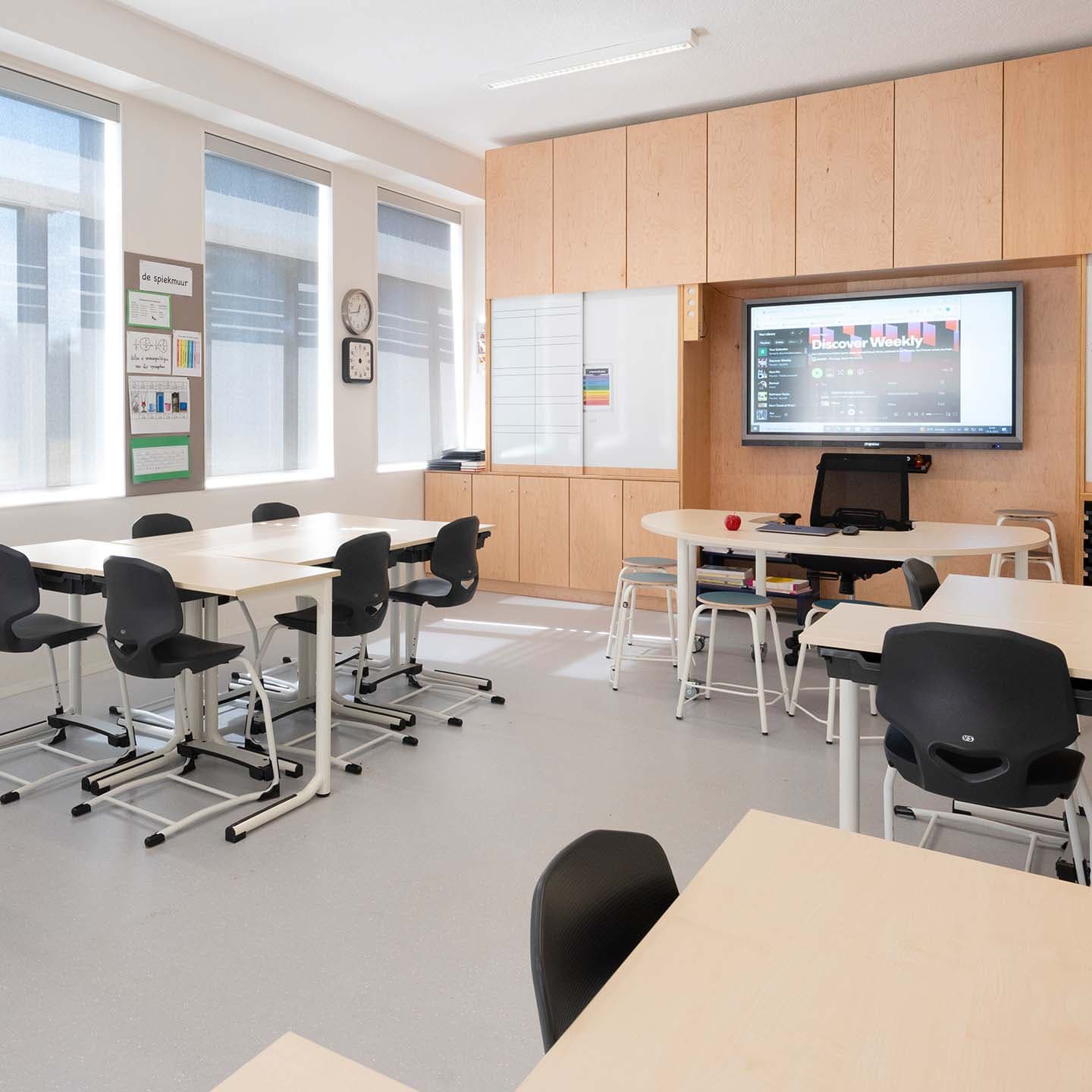Modern klaslokaal met witte tafels, zwarte stoelen, een digitaal scherm, grote ramen en houten opbergkasten.