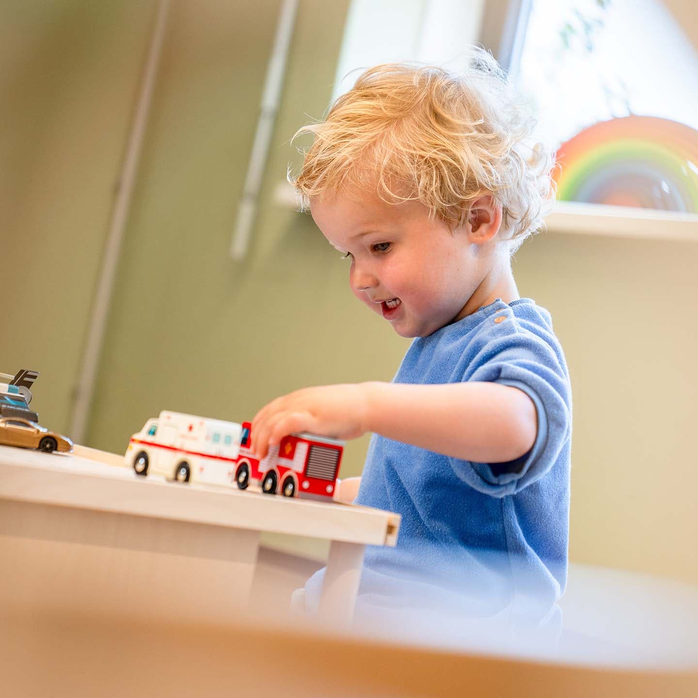 Een jong kind met krullend haar speelt met speelgoedvrachtwagens op een tafel in een lichte kamer, met een regenboogdecoratie op de achtergrond. Zonlicht stroomt naar binnen door een groot raam.