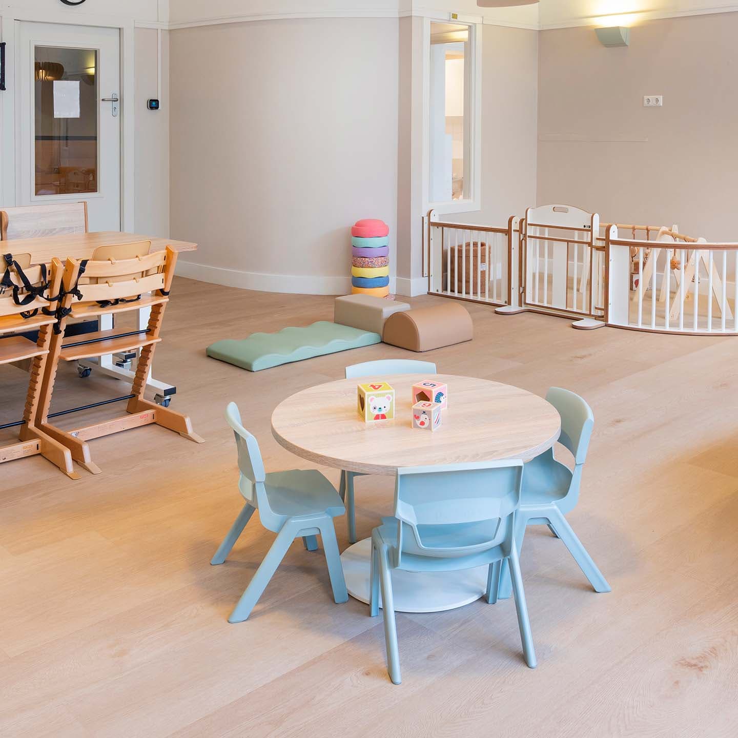 Neutraal ingerichte babygroep in de kinderopvang met een kleine ronde kindertafel, lichtgroene stoelen, een houten grondbox en softplay speelmatten.
