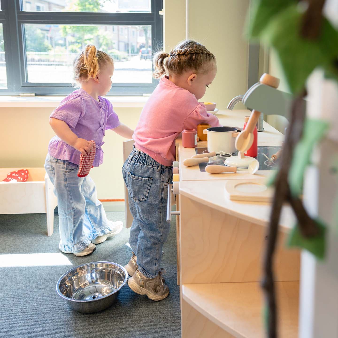 Twee kinderen spelen in een speelkeuken; de ene roert in een pan, de andere houdt een doek vast. Zonlicht stroomt naar binnen door een groot raam.