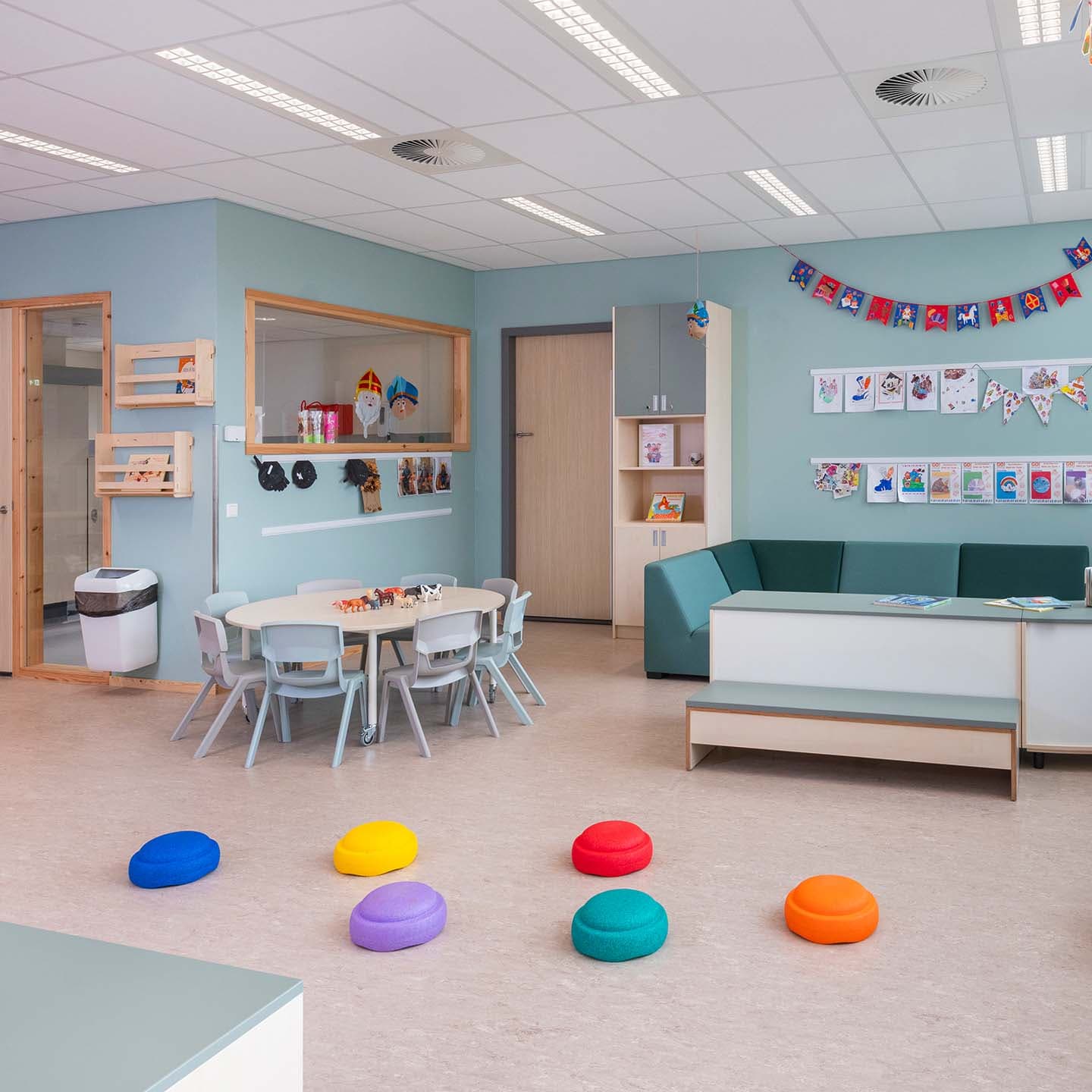 Een lichte en kleurrijke groepsruimte in een kinderdagverblijf met kleine tafels, stoelen, kussens en kunstwerken aan de muren. Ook zijn planken en educatieve materialen zichtbaar.