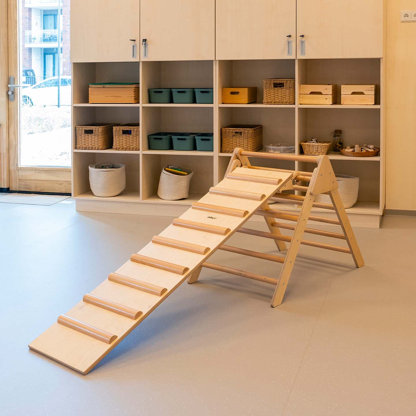 Houten klimtoestel met hellingbaan in een groepsruimte met houten open opbergkasten waarin opbergbakken geplaatst zijn.