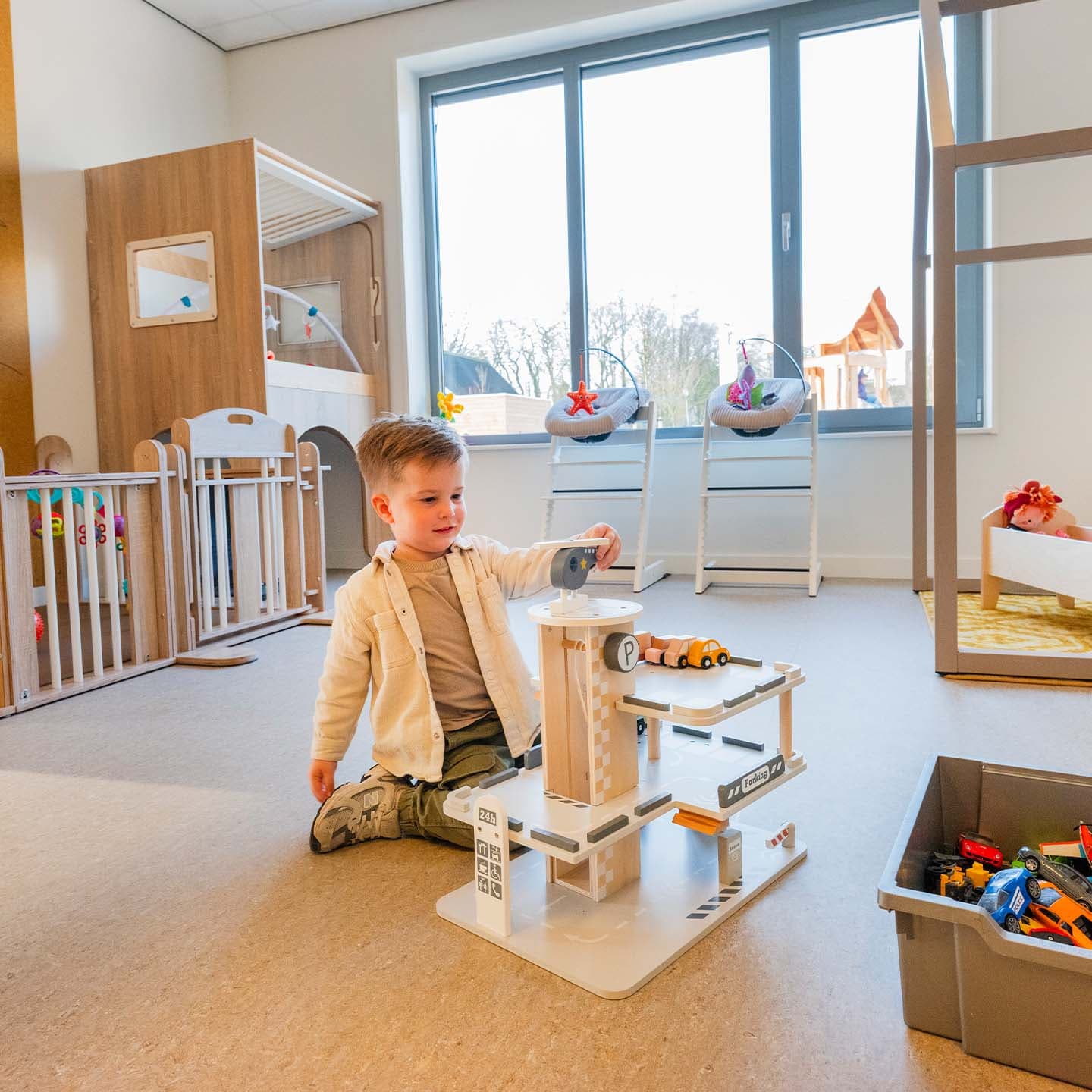 Kind speelt met een speelgoedgarage op de vloer in een lichte groepsruimte met spelmateriaal en grote ramen.