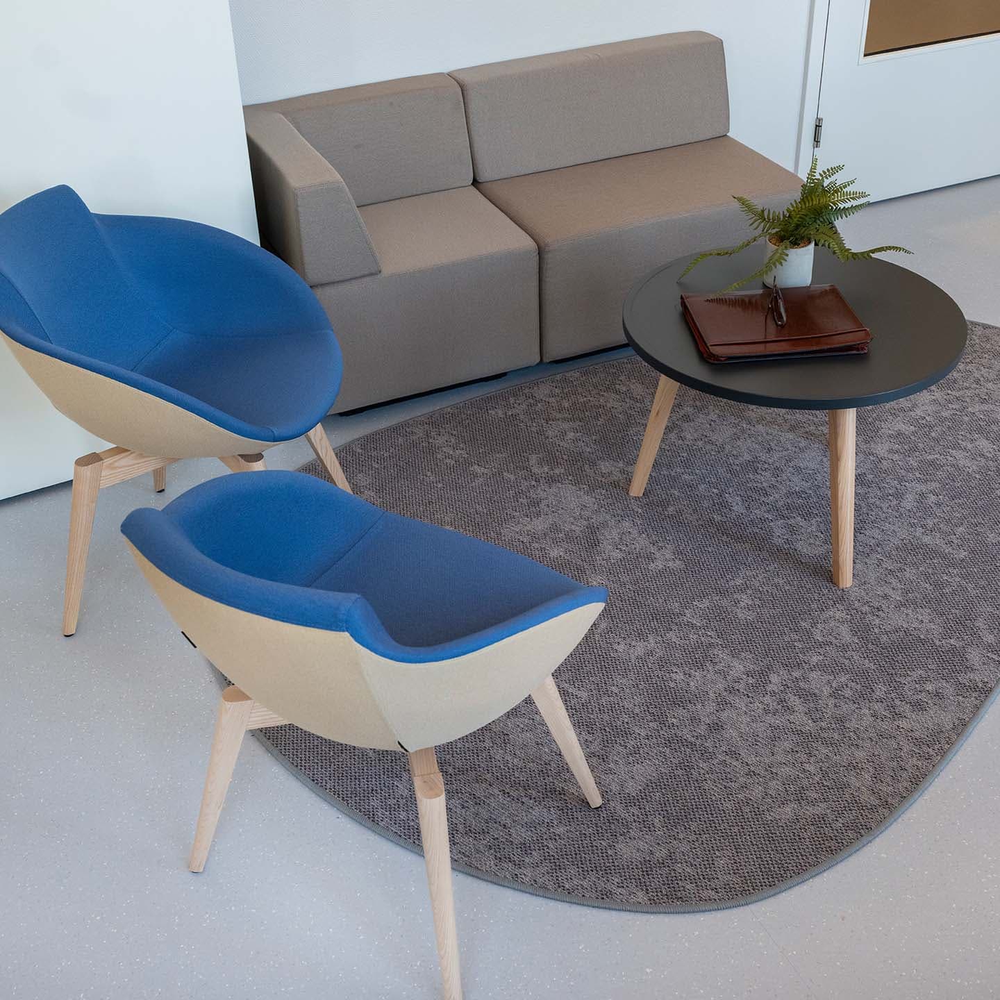 Moderne zithoek met twee blauwe stoelen, een grijze bank, een ronde tafel met boeken en een plant, op een grijs vloerkleed.
