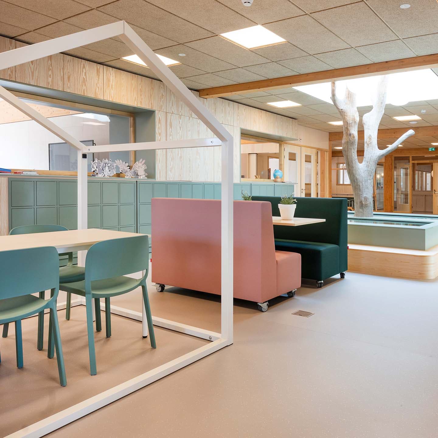 Open ruimte met een tafel onder een huisvormige constructie, gekleurde banken en opbergkasten op kinderhoogte.