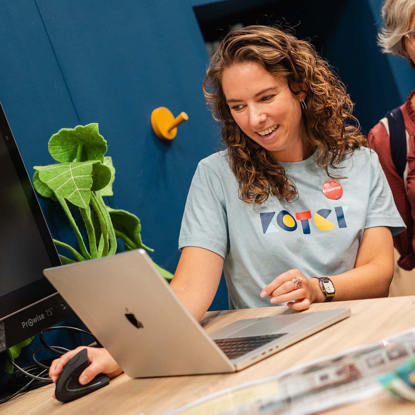 Een lachende vrouw met een Koter-shirt en een laptop met groene plant op de achtergrond