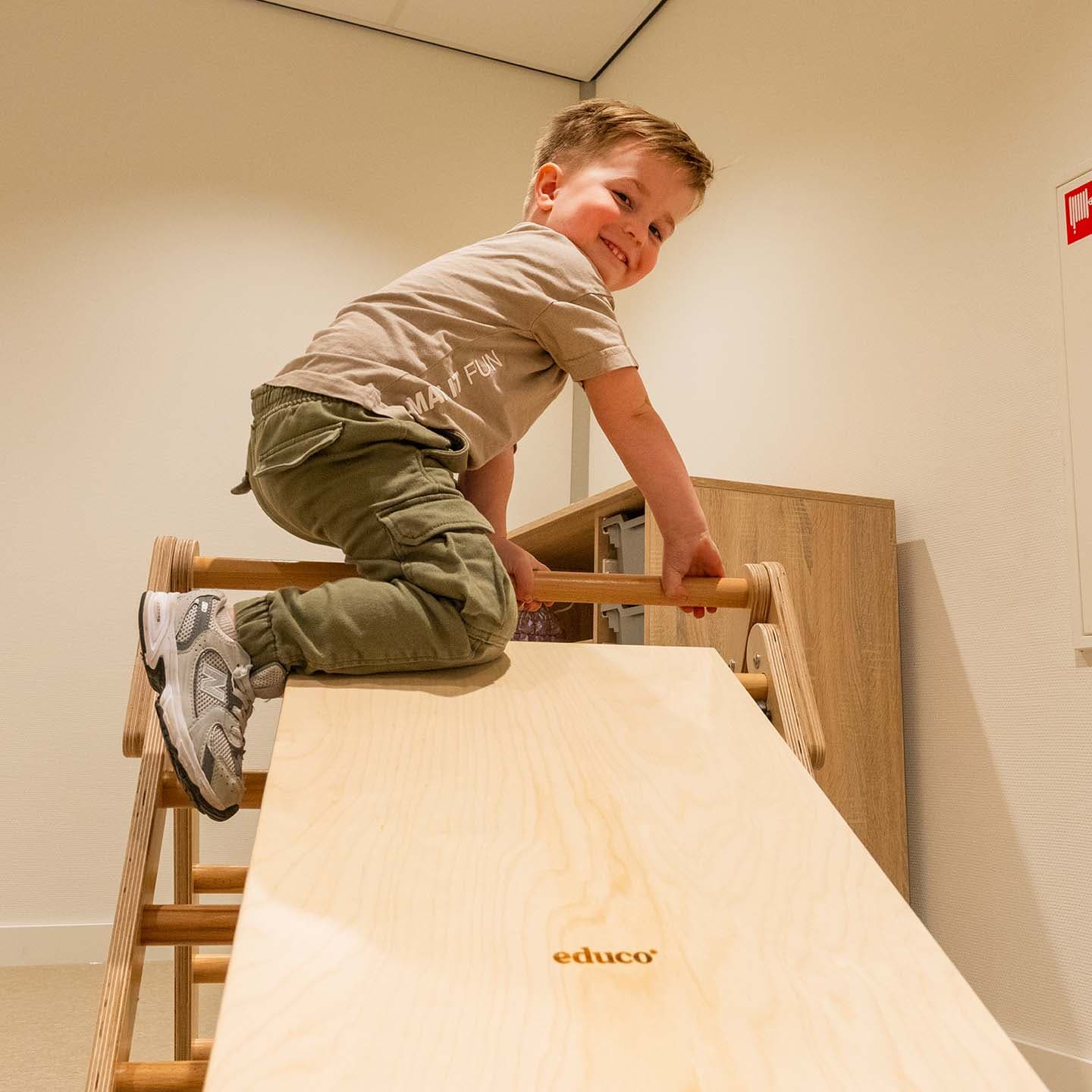 Een kleine jongen klimt op een houten klimdriehoek met een glijplank in de kinderopvang.