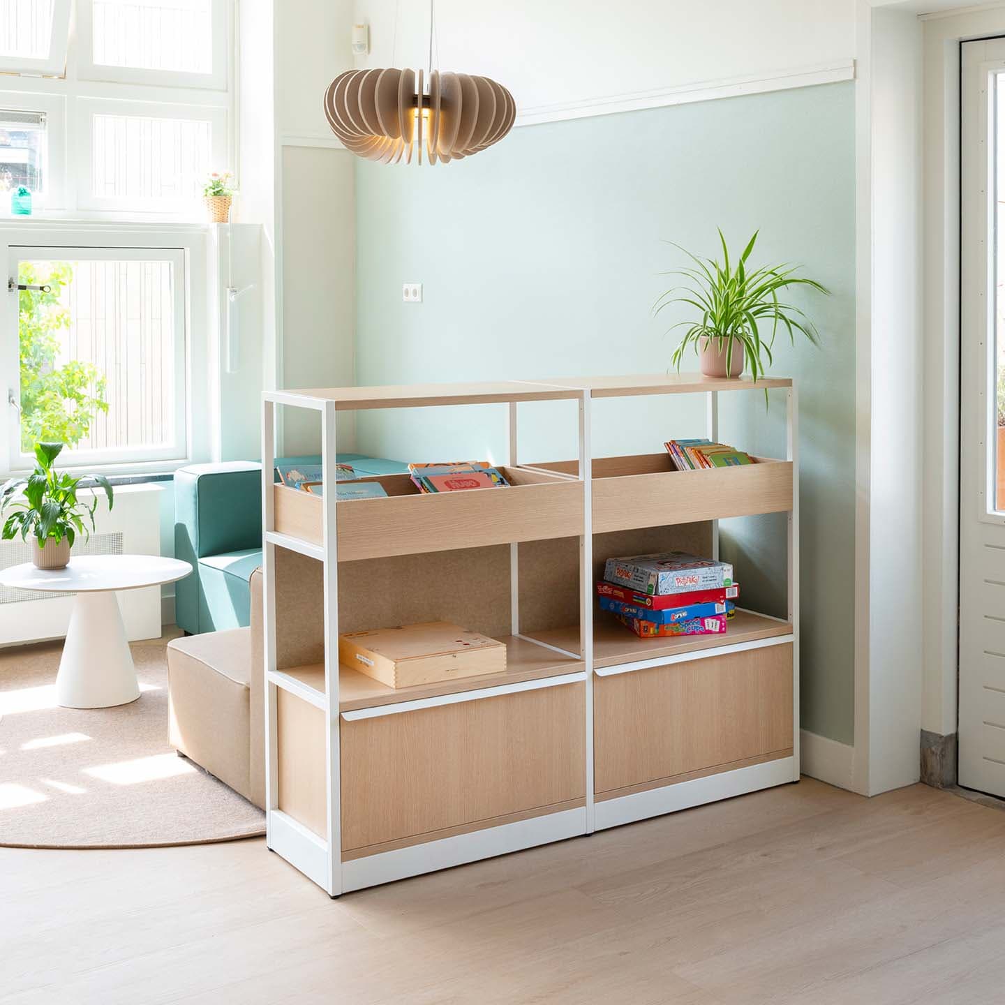 Groepsruimte in de kinderopvang met een moderne houten boekenkast met houten opbergbakken met daarachter een comfortabele groene bank bij het raam.