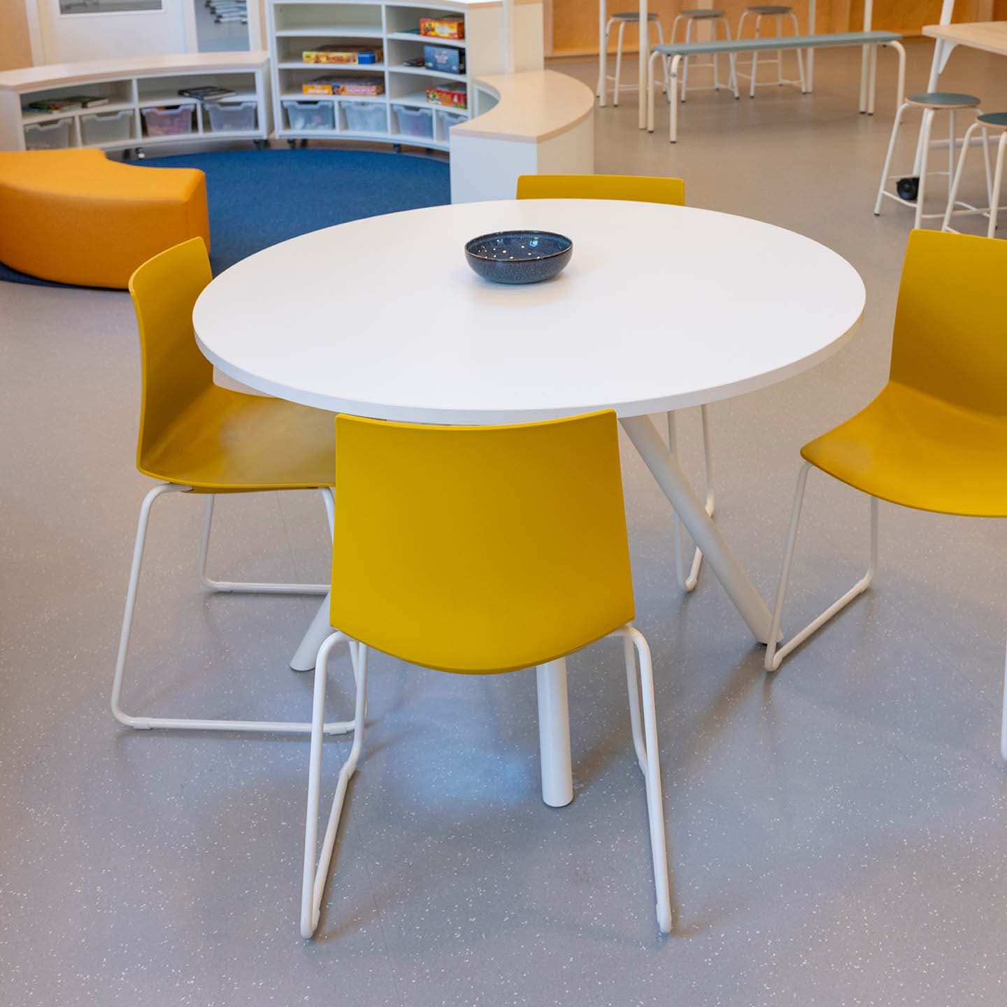 Groepsruimte op de BSO met een ronde witte tafel met een schaal, omringd door vier gele stoelen in een lichte ruimte met boekenkasten en extra zitplekken op de achtergrond.