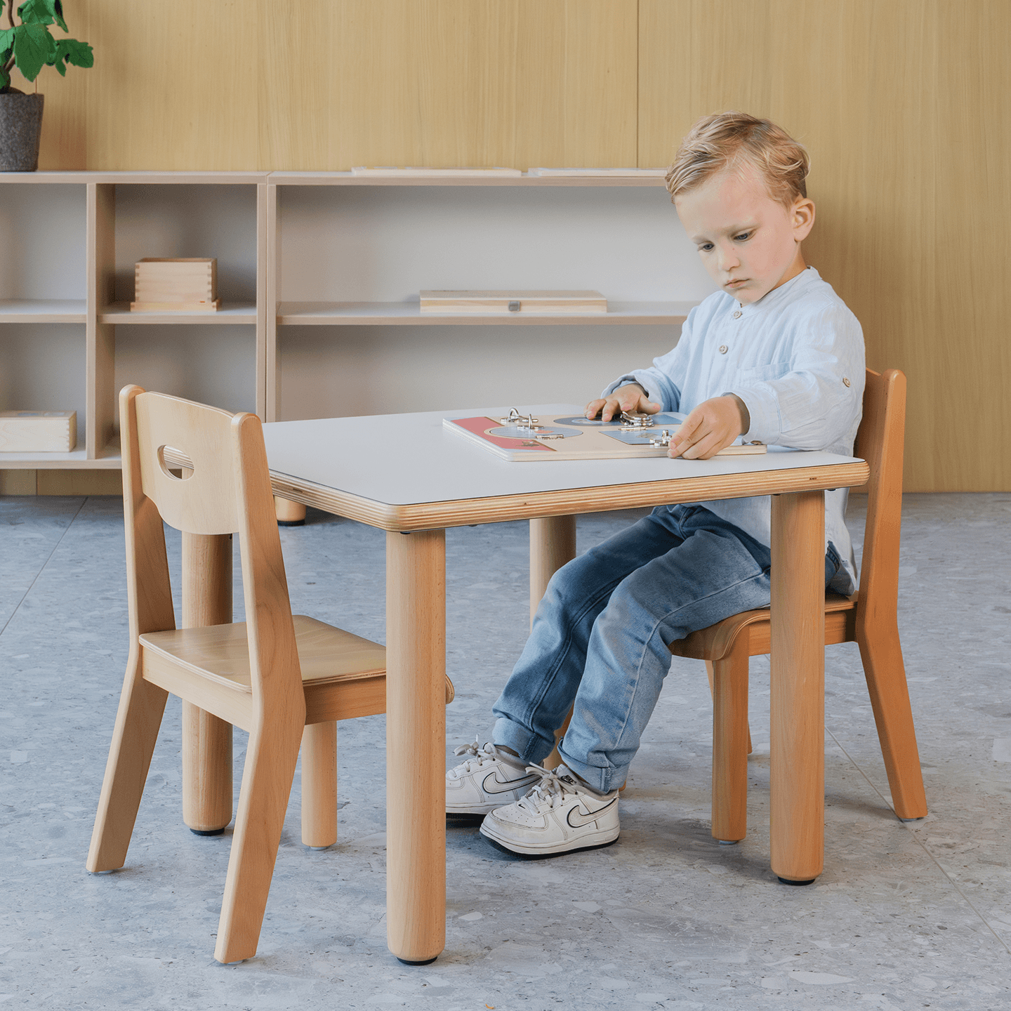 Een jong kind zit aan een houten tafel in een kinderdagverblijf en speelt en ontwikkelt hiermee de fijne motoriek. Op de achtergrond zijn planken en een plant zichtbaar.