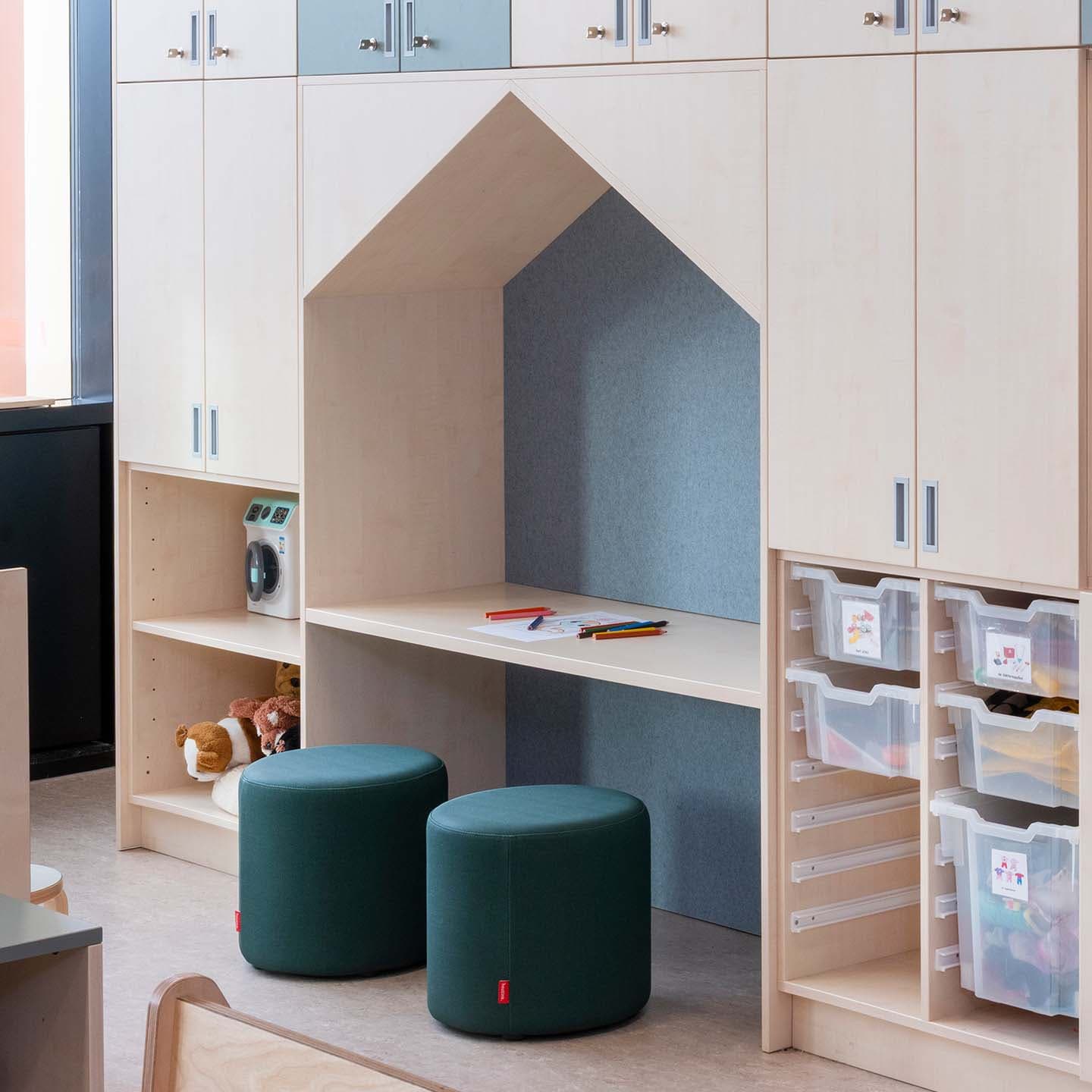 Op maat gemaakte houten wandkast met een ingebouwde huisvormige werkplek waar poefjes onderstaan. De kast heeft open en dichte vakken met plastic opbergbakken.