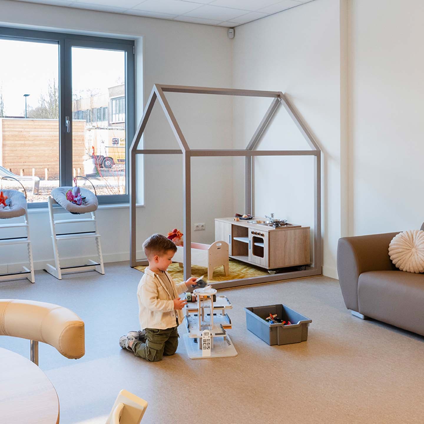 Een kind speelt met speelgoed op de vloer in een lichte ruimte van een kinderdagverblijf. Op de achtergrond staan een kleine speelhuisconstructie en een speelgoedkeuken bij grote ramen.