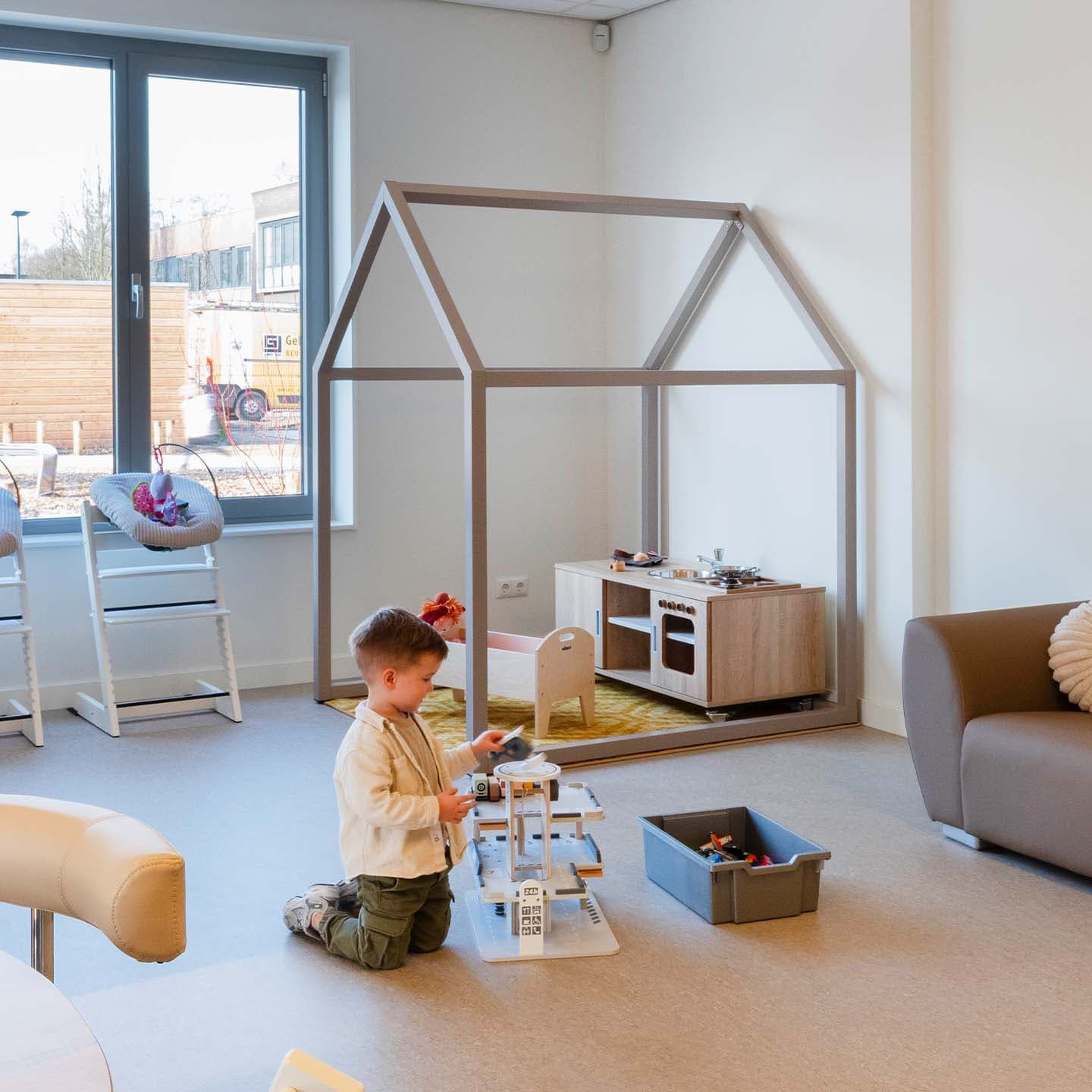 Een kleine jongen speelt met een autogarage op de vloer in de kinderopvang met een speelhuisje en kinderkeuken op de achtergrond.