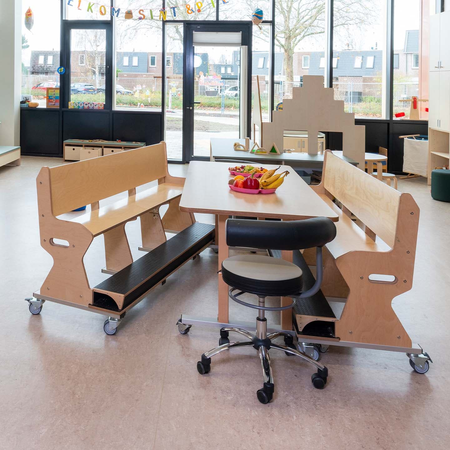 Kinderopvang groepsruimte met een verrijdbare houten tafel, houten ergobanken op wieltjes en een leidsterstoel.
