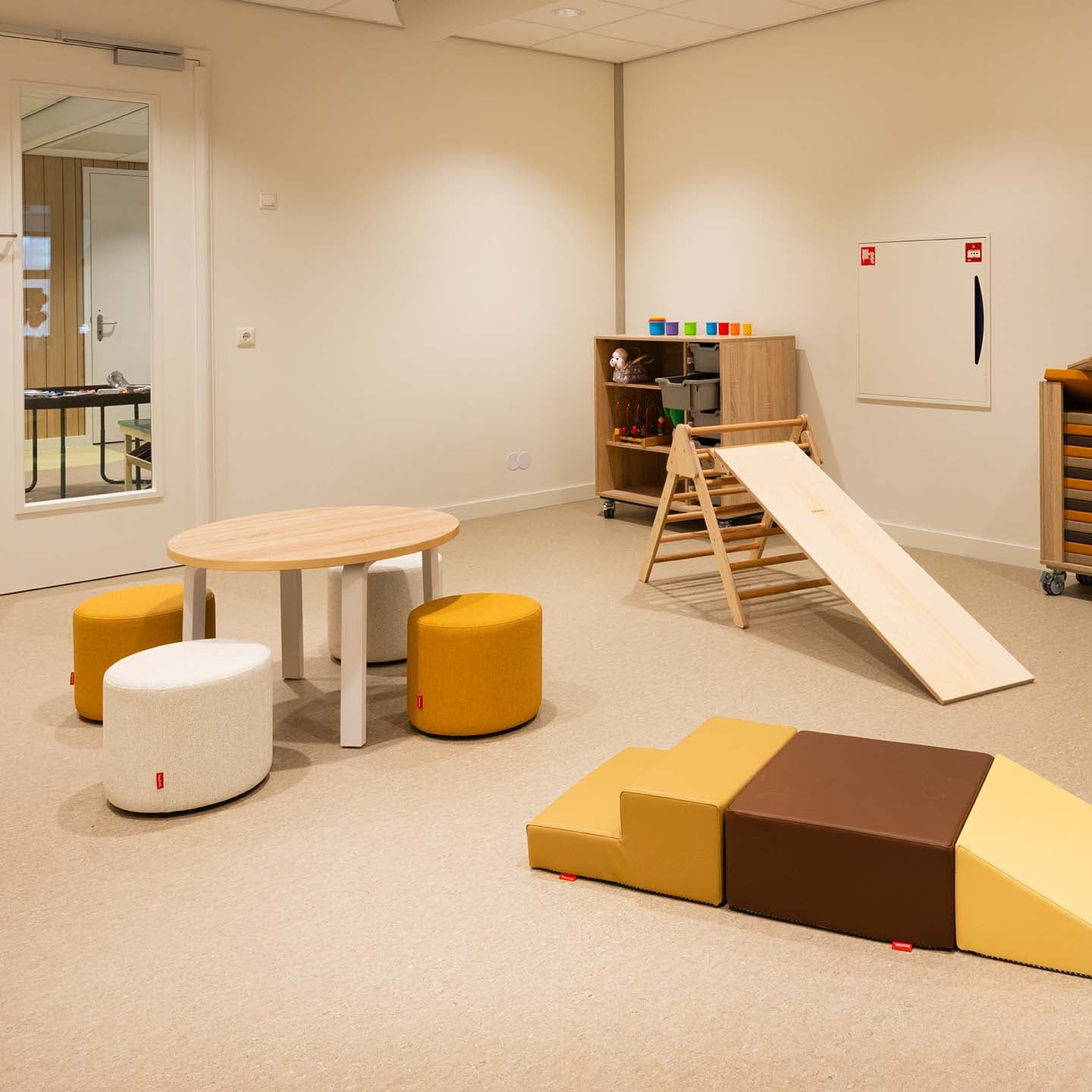 Speelruimte in de kinderopvang met een kleine kindertafel, ronde krukjes, een houten klimrek met glijbaan, softplay speelelementen en een opbergkast.