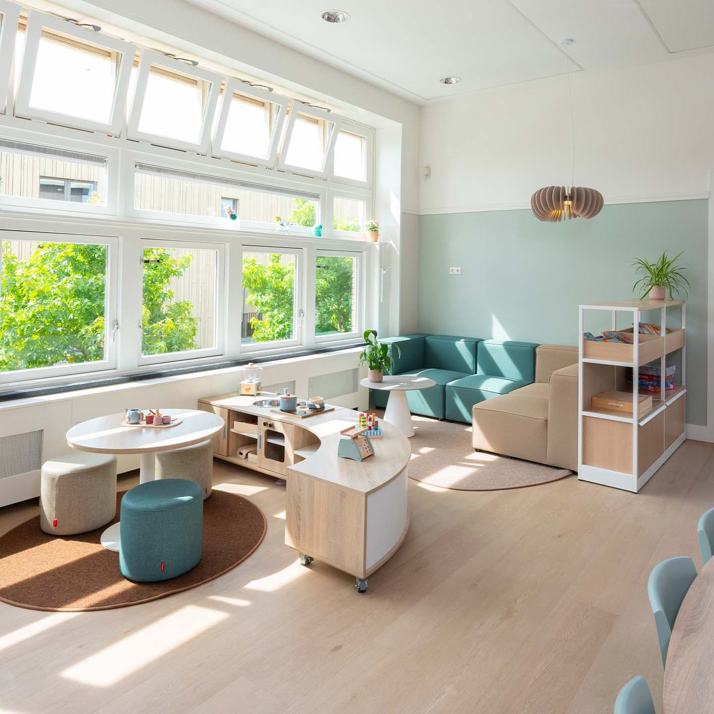Een lichte kinderopvang met moderne meubels, waaronder tafels, poefs en een bank bij grote ramen. Op de planken staan boeken en een plant. Natuurlijk licht vult de ruimte.