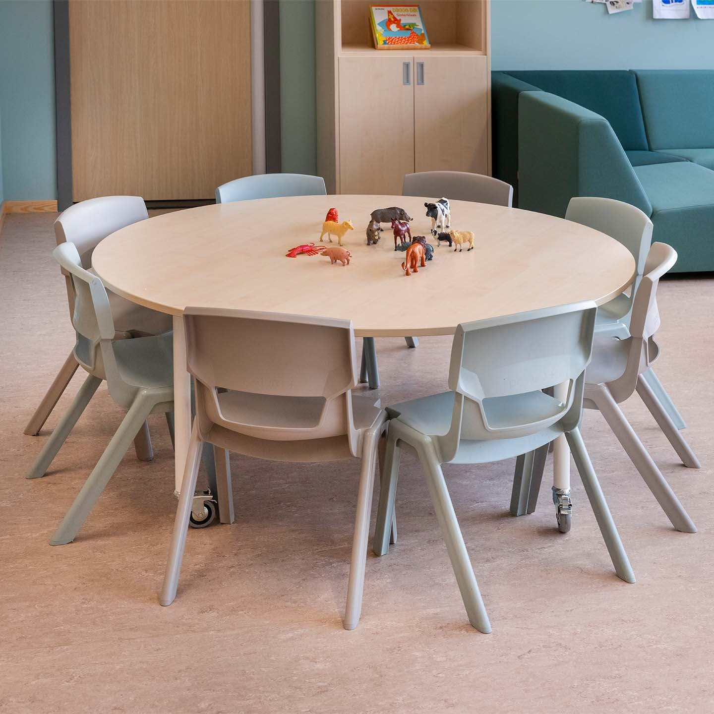 Groepsruimte in de kinderopvang met een ronde kindertafel, zes stoelen, speelgoeddieren op tafel, een bank en een boekenkast.