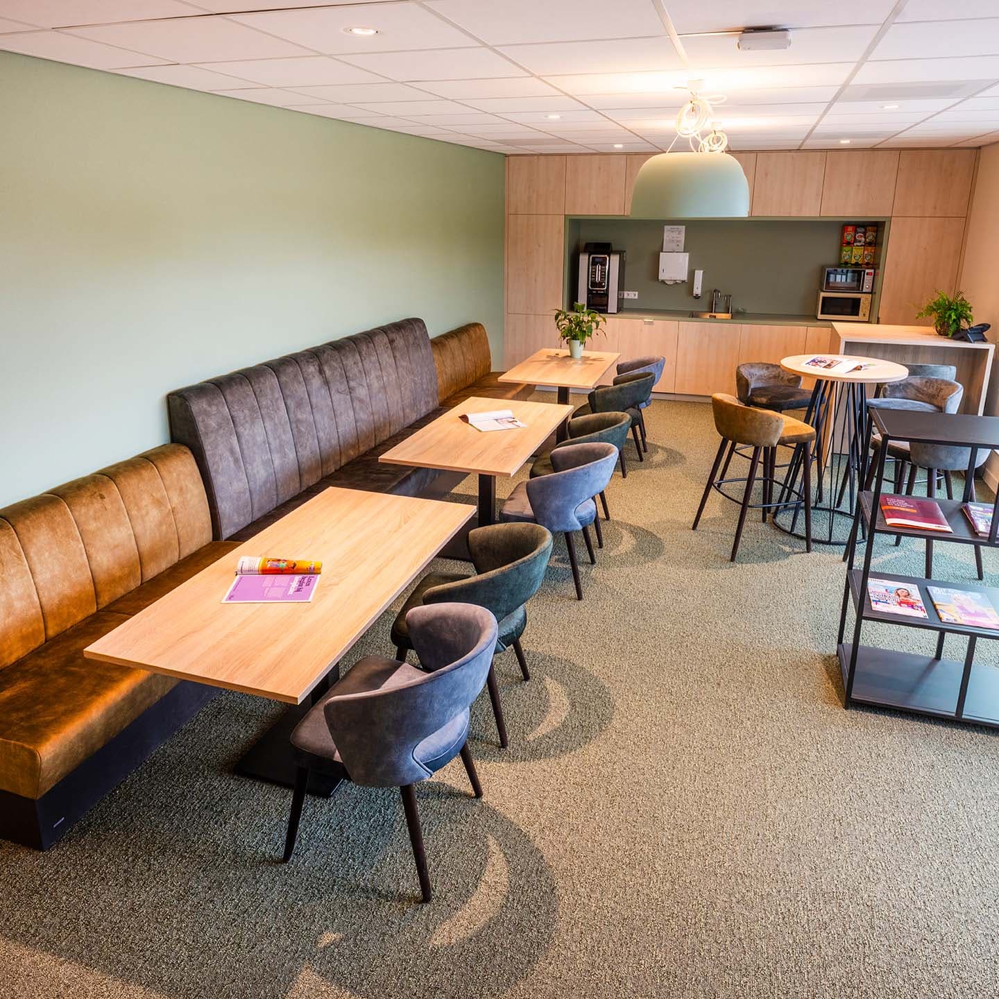Een ruime teamkamer met comfortabele zitplekken, wandbanken, een koffiecorner en robuuste houten tafels. Modern lounge with light wood tables, brown bench seating, gray chairs, coffee station, magazines, and green accent wall.