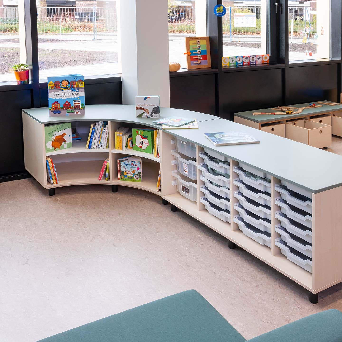 Lees- en speelhoek in de kinderopvang met een lage, gebogen ruimteverdeler die fungeert als boekenkast en opbergplek.