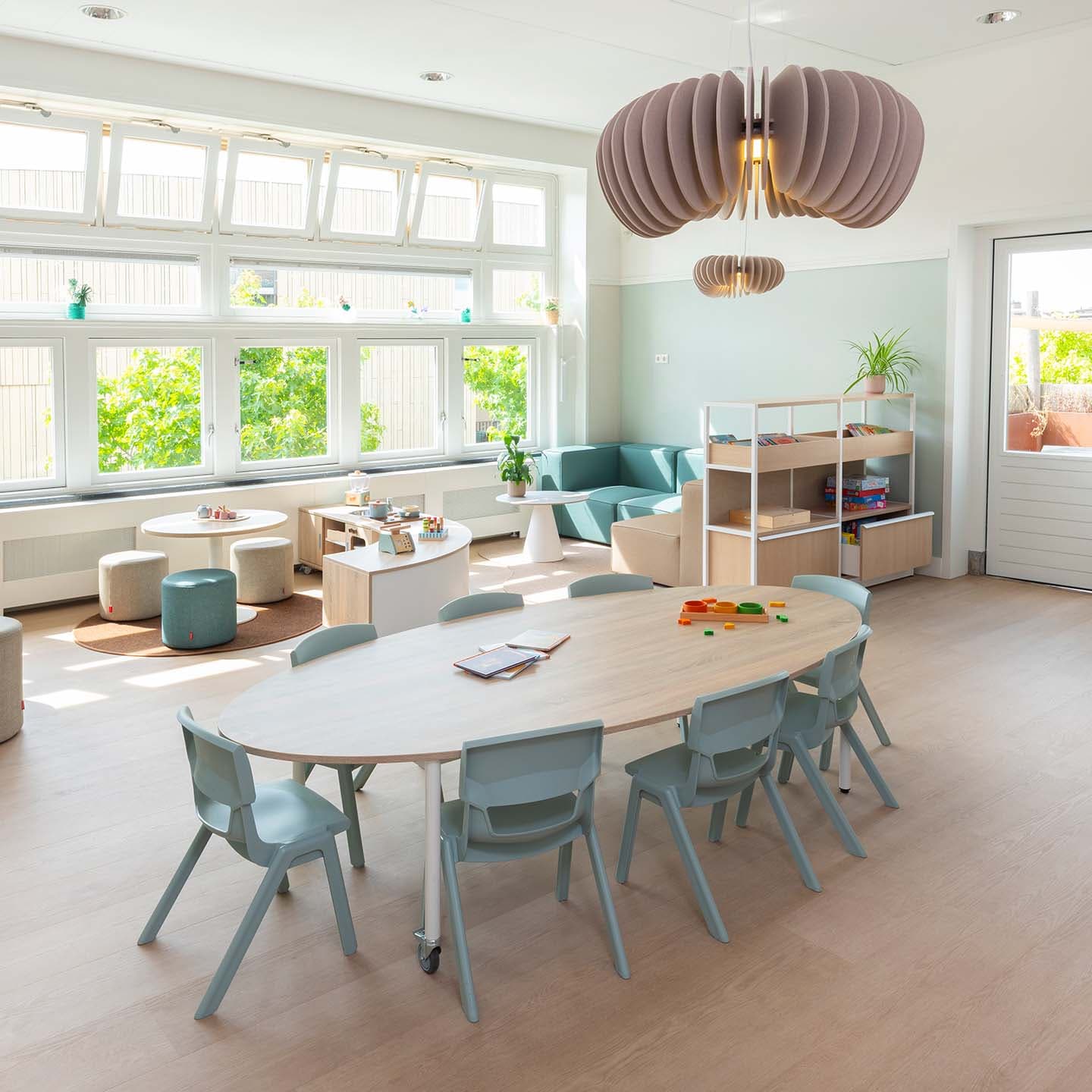 Een lichte, moderne speelruimte in een kinderdagverblijf met ronde tafels, zachte zitplekken, speelgoed en boekenkasten. Grote ramen met uitzicht op groene bomen laten natuurlijk licht binnen.