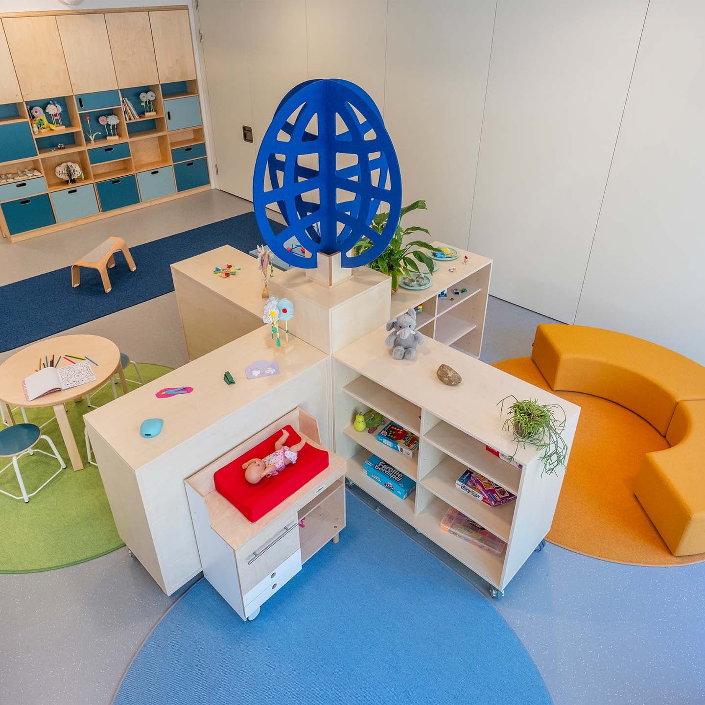 Groepsruimte in de kinderopvang met een blauwe speelboom, opbergkasten op kinderhoogte in een X-opstelling en gebogen oranje zitbanken.